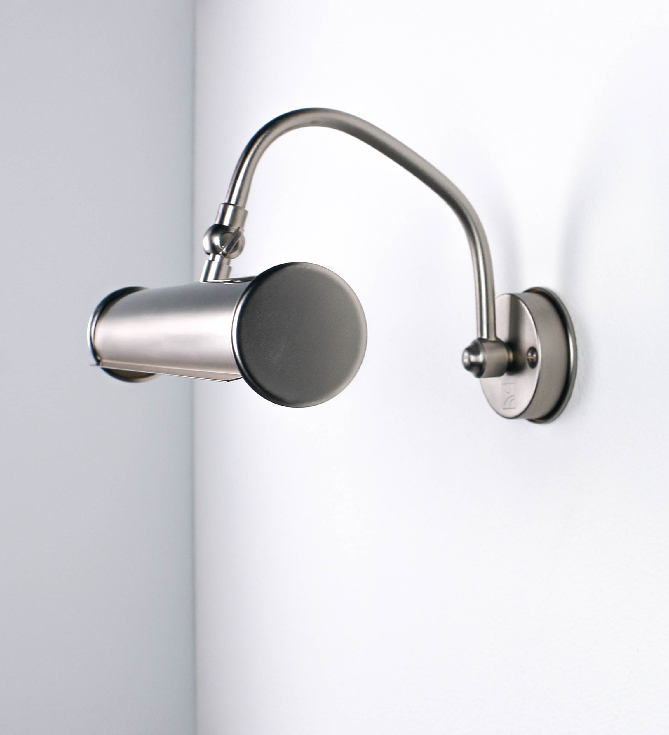 Aplique de parede que combina elementos clássicos com toques contemporâneos, todo em níquel escovado. O aplique é composto por uma base circular de onde se estende um braço curvo e gracioso, que suporta a barra principal. O braço contém um elemento que permite apertar ou desapertar a barra para melhor manipular a direção da luz. A barra de 25cm de largura é cilíndrica e alongada. As extremidades do cilindro são planas e do mesmo diâmetro.