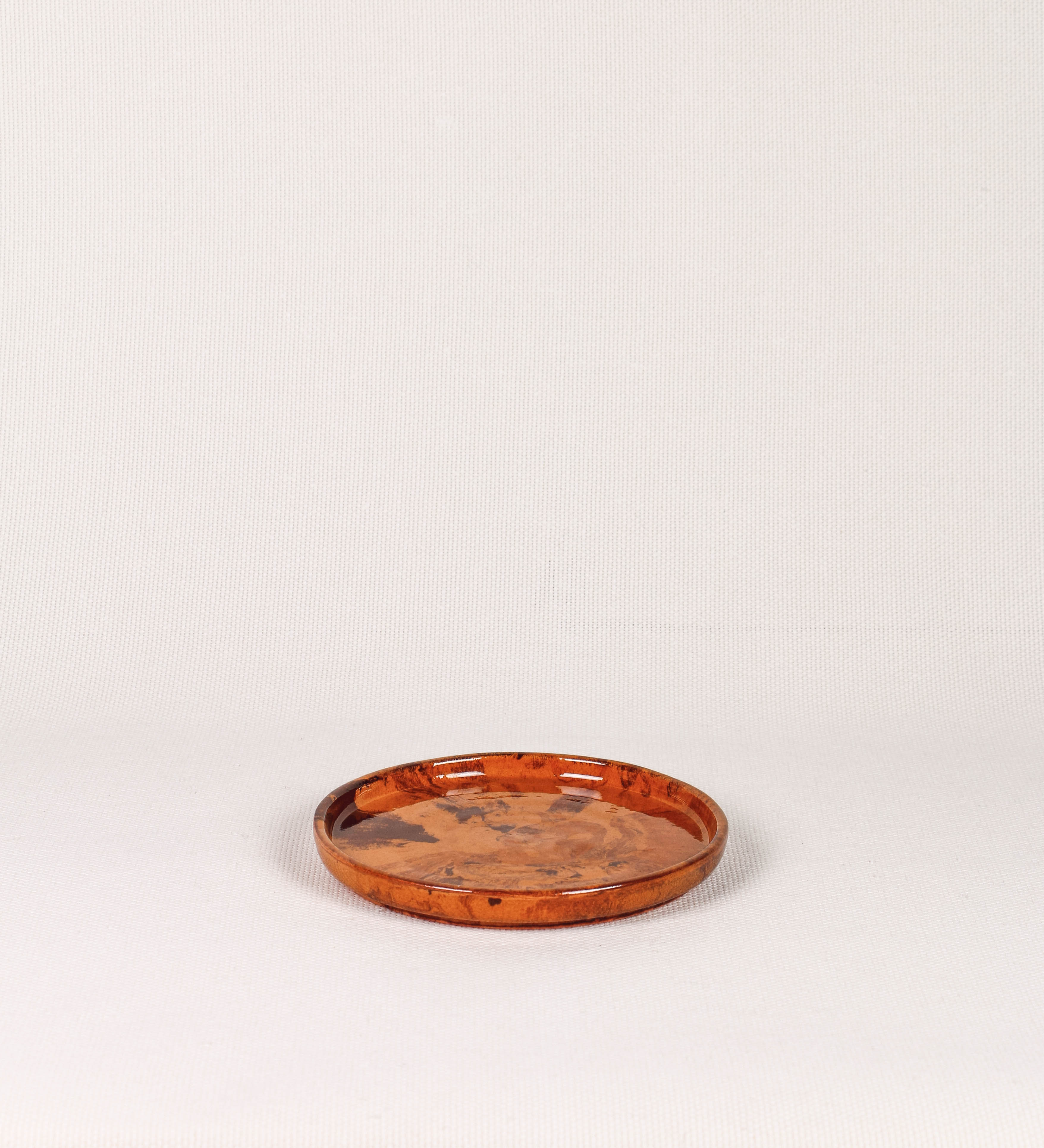 Prato de sobremesa com design minimalista e distinto. O prato é de barro, com uma mistura de dois tipos de barro diferentes, um mais escuro e outro mais claro. Esta mistura cria um desenho de formas abstratas e aleatórias, que se espalham por todas as superfícies do prato. Tem acabamento vidrado castanho translúcido, escurecendo as cores originais dos barros misturados. A forma do prato é circular, com uma borda envolvente de altura baixa e design simples e minimalista.