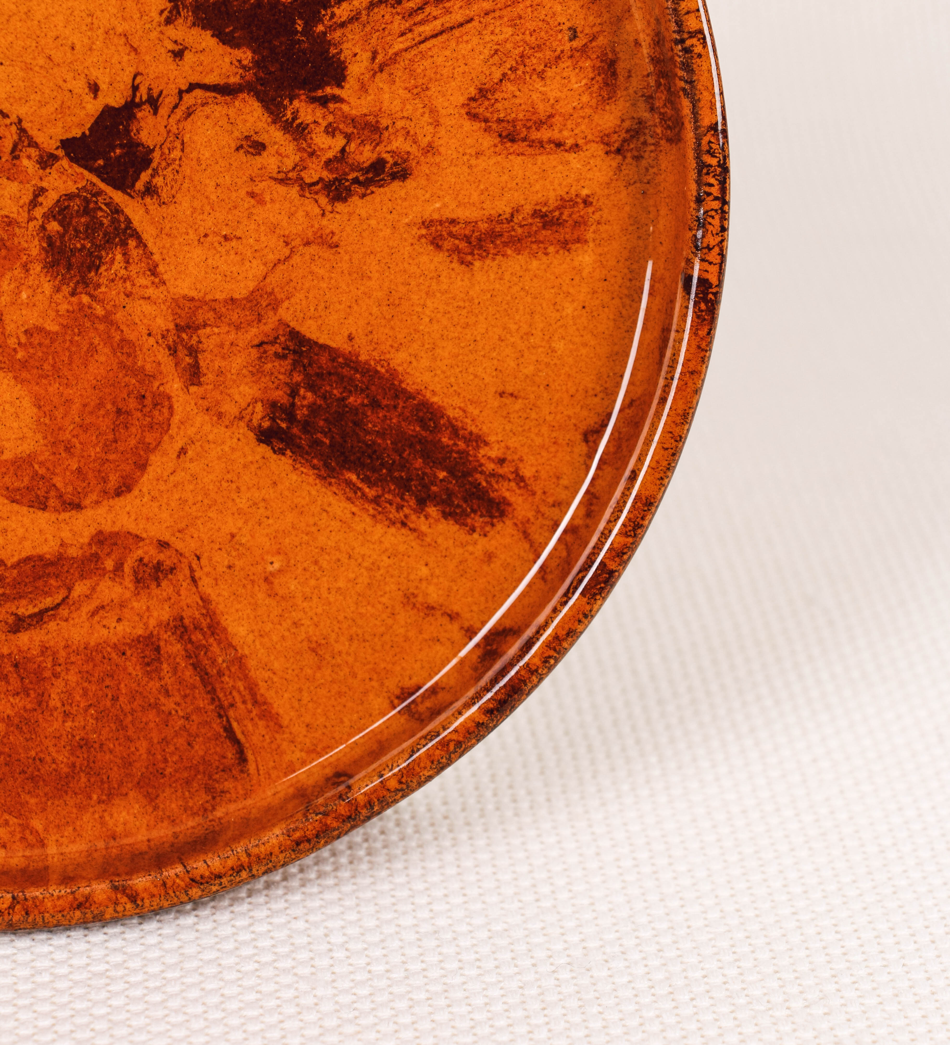 Prato de sobremesa com design minimalista e distinto. O prato é de barro, com uma mistura de dois tipos de barro diferentes, um mais escuro e outro mais claro. Esta mistura cria um desenho de formas abstratas e aleatórias, que se espalham por todas as superfícies do prato. Tem acabamento vidrado castanho translúcido, escurecendo as cores originais dos barros misturados. A forma do prato é circular, com uma borda envolvente de altura baixa e design simples e minimalista.