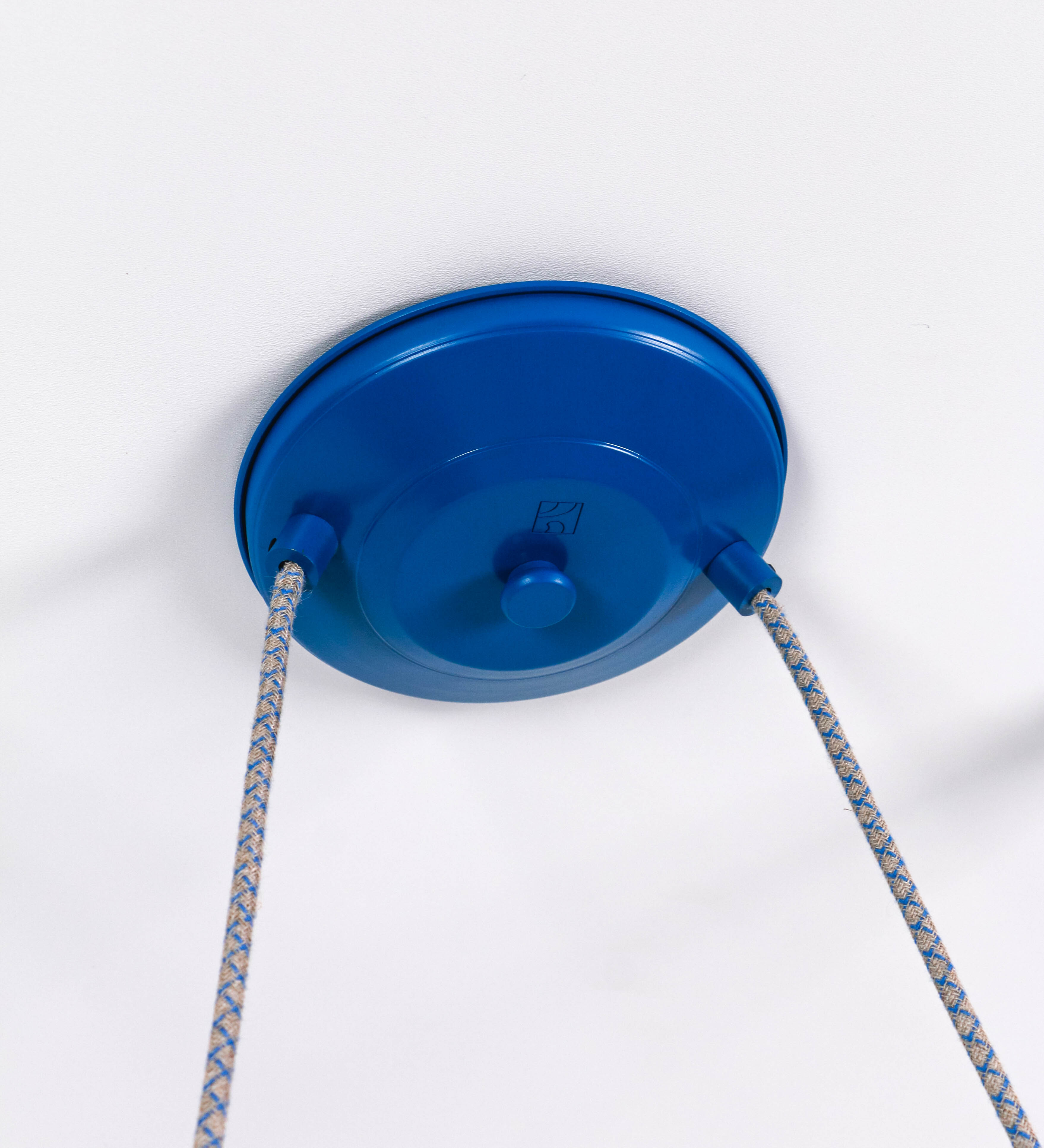 Suspensão de teto dupla composta por uma base circular azul vintage de onde descem dois cabos revestidos têxtil com linhas azuis e beges. Cada cabo passa por uma roldana em latão escovado fixa ao teto e desce até aos chapéus. Entre a base e as roldanas cada cabo sustenta uma esfera em latão escovado que atua como contrapeso para ajustar a altura. Os dois chapéus são azuis de formato cónico invertido e interior branco. No topo de cada chapéu há um suporte em latão escovado que conecta os chapéus aos cabos.