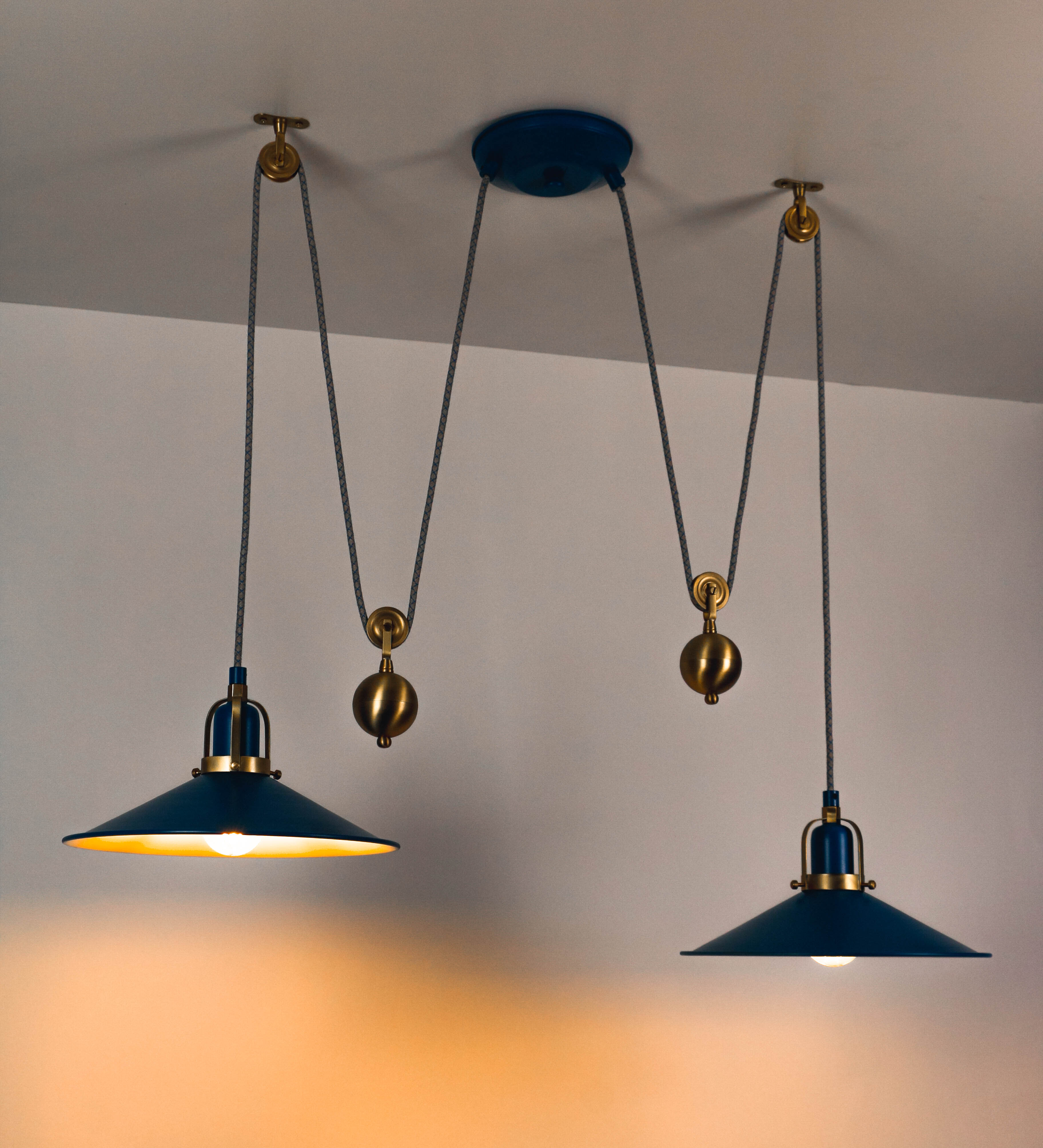 Suspensão de teto dupla composta por uma base circular azul vintage de onde descem dois cabos revestidos têxtil com linhas azuis e beges. Cada cabo passa por uma roldana em latão escovado fixa ao teto e desce até aos chapéus. Entre a base e as roldanas cada cabo sustenta uma esfera em latão escovado que atua como contrapeso para ajustar a altura. Os dois chapéus são azuis de formato cónico invertido e interior branco. No topo de cada chapéu há um suporte em latão escovado que conecta os chapéus aos cabos.