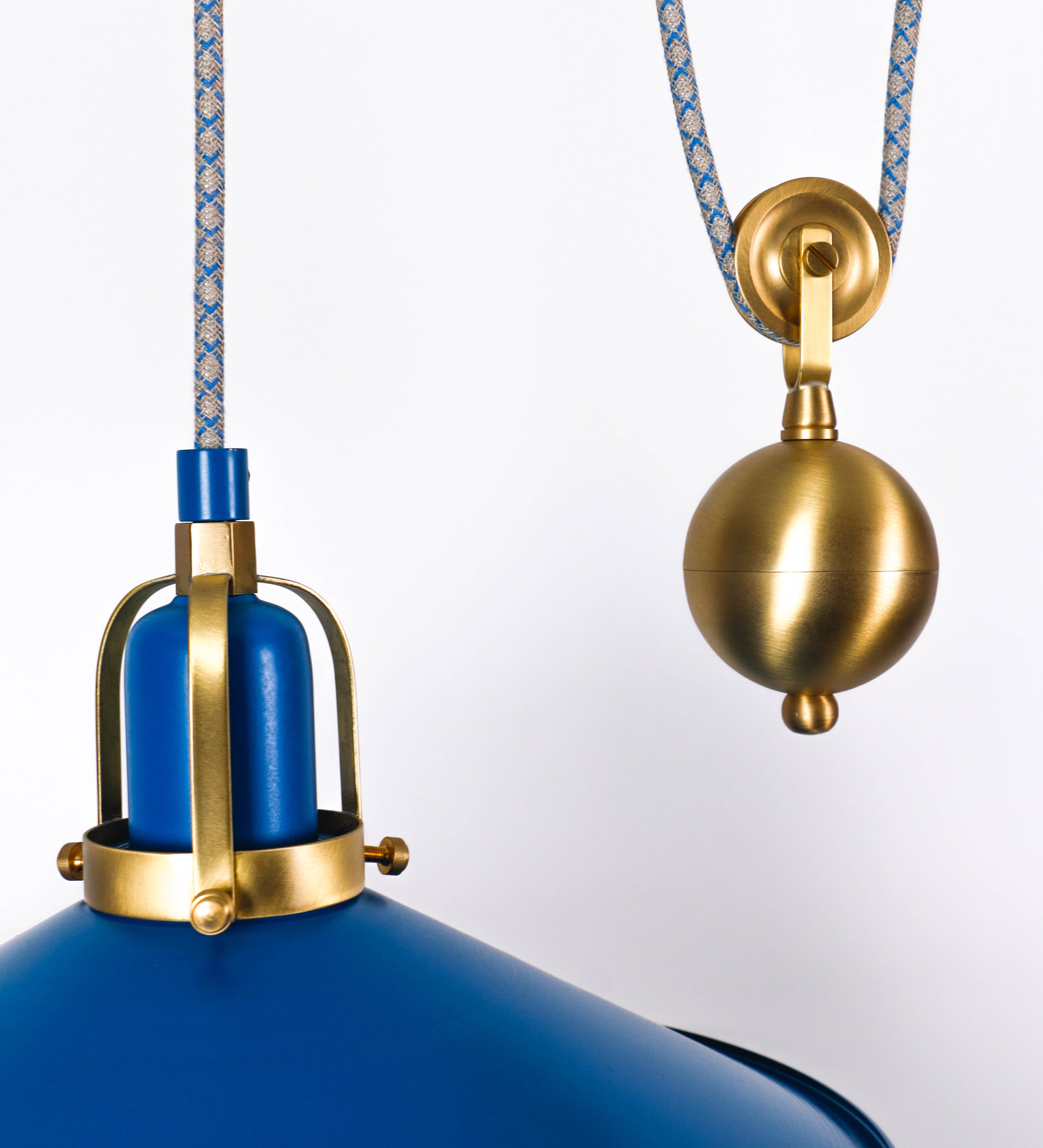 Suspensão de teto dupla composta por uma base circular azul vintage de onde descem dois cabos revestidos têxtil com linhas azuis e beges. Cada cabo passa por uma roldana em latão escovado fixa ao teto e desce até aos chapéus. Entre a base e as roldanas cada cabo sustenta uma esfera em latão escovado que atua como contrapeso para ajustar a altura. Os dois chapéus são azuis de formato cónico invertido e interior branco. No topo de cada chapéu há um suporte em latão escovado que conecta os chapéus aos cabos.