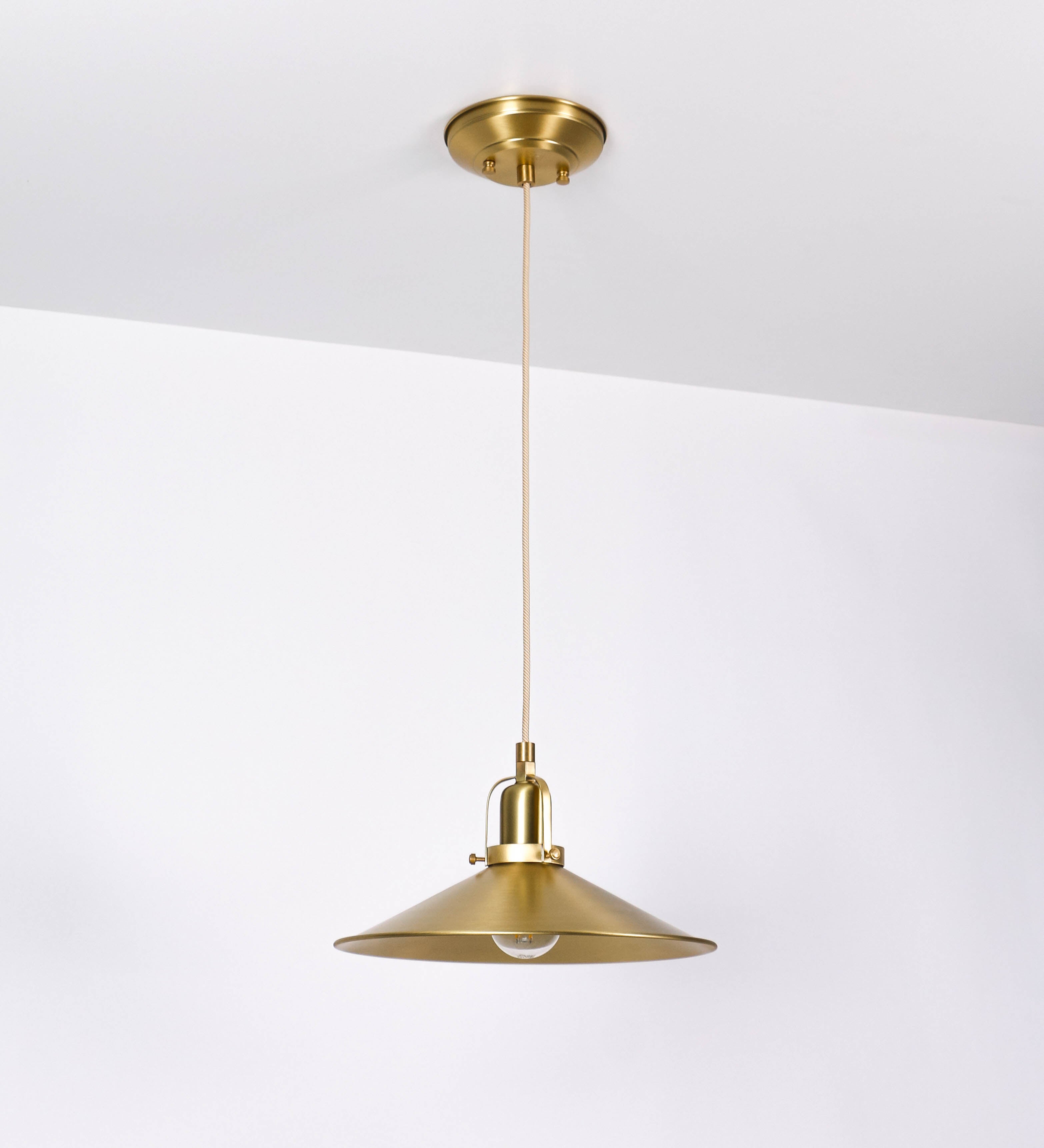 Suspensão de teto que combina um design clássico com um acabamento moderno e sofisticado. A suspensão, toda em latão escovado, é fixa ao teto através de uma base circular de onde desce um cabo revestido têxtil com um padrão trançado com linhas douradas e brancas, que sustenta o chapéu principal. O chapéu tem um formato cónico invertido, com uma borda ligeiramente levantada. No topo do chapéu, há um suporte circular com três pequenos braços que conecta o chapéu ao cabo.