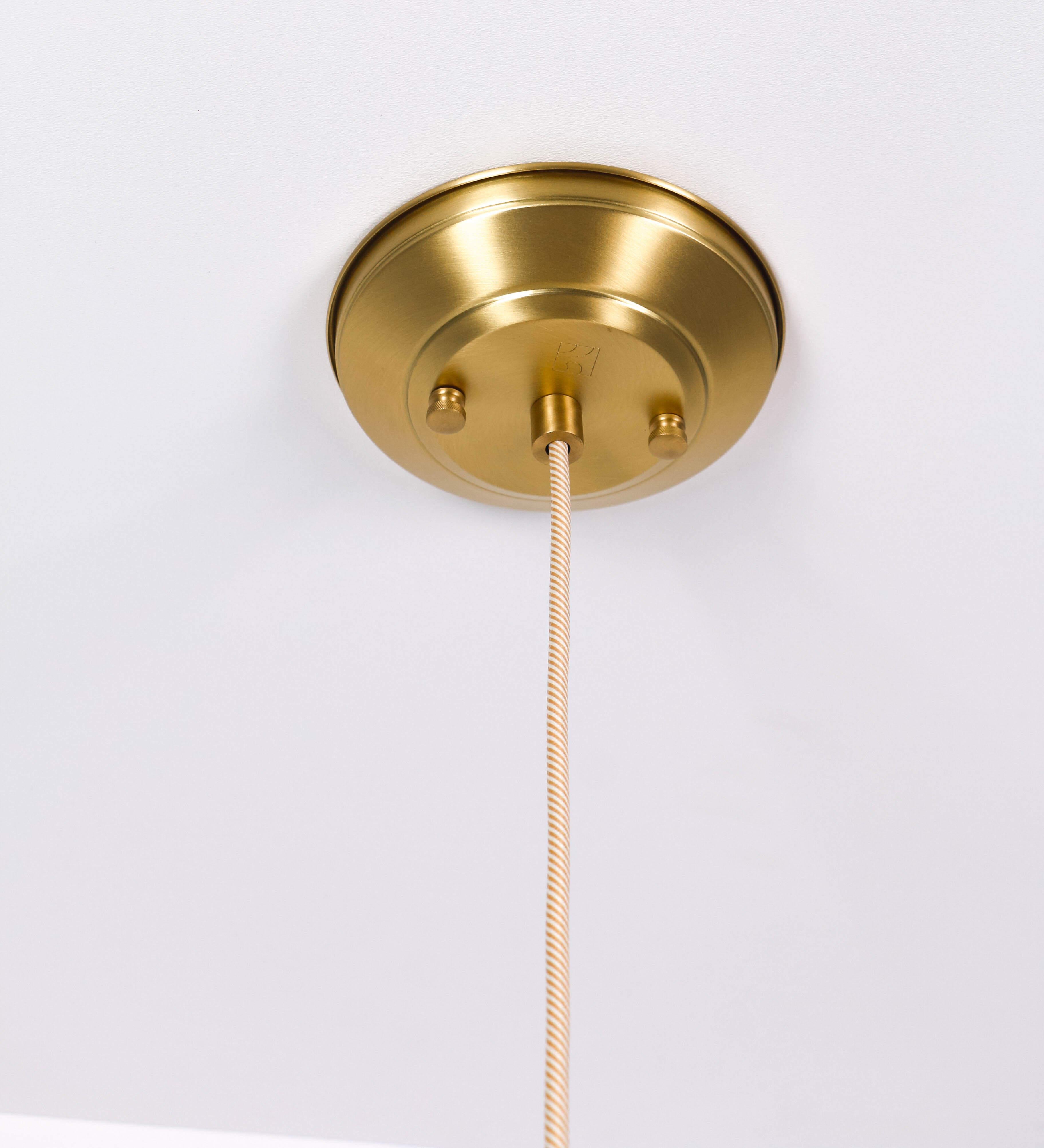 Suspensão de teto que combina um design clássico com um acabamento moderno e sofisticado. A suspensão, toda em latão escovado, é fixa ao teto através de uma base circular de onde desce um cabo revestido têxtil com um padrão trançado com linhas douradas e brancas, que sustenta o chapéu principal. O chapéu tem um formato cónico invertido, com uma borda ligeiramente levantada. No topo do chapéu, há um suporte circular com três pequenos braços que conecta o chapéu ao cabo.