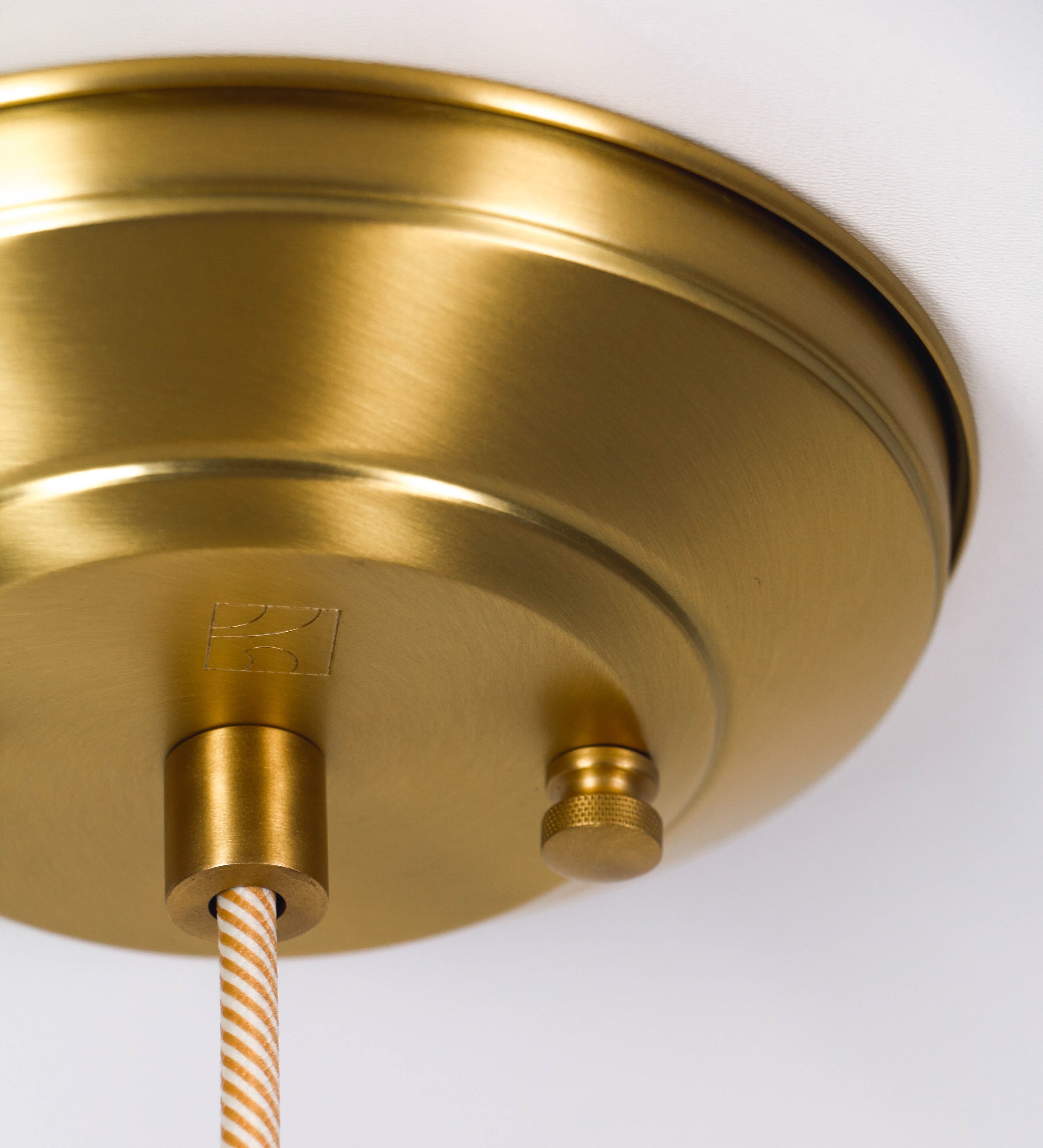 Suspensão de teto que combina um design clássico com um acabamento moderno e sofisticado. A suspensão, toda em latão escovado, é fixa ao teto através de uma base circular de onde desce um cabo revestido têxtil com um padrão trançado com linhas douradas e brancas, que sustenta o chapéu principal. O chapéu tem um formato cónico invertido, com uma borda ligeiramente levantada. No topo do chapéu, há um suporte circular com três pequenos braços que conecta o chapéu ao cabo.