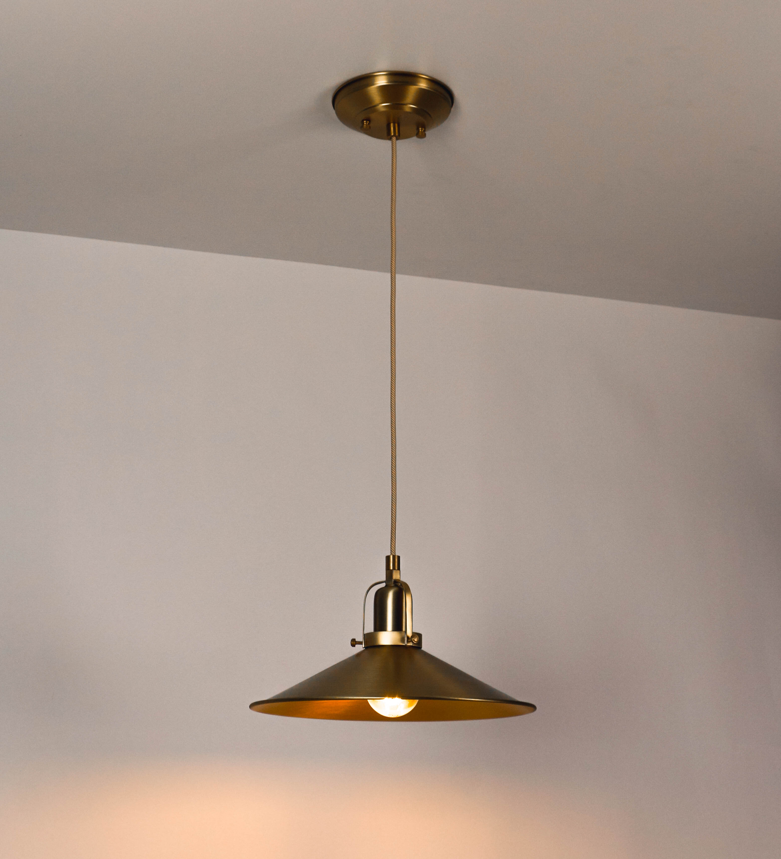 Suspensão de teto que combina um design clássico com um acabamento moderno e sofisticado. A suspensão, toda em latão escovado, é fixa ao teto através de uma base circular de onde desce um cabo revestido têxtil com um padrão trançado com linhas douradas e brancas, que sustenta o chapéu principal. O chapéu tem um formato cónico invertido, com uma borda ligeiramente levantada. No topo do chapéu, há um suporte circular com três pequenos braços que conecta o chapéu ao cabo.