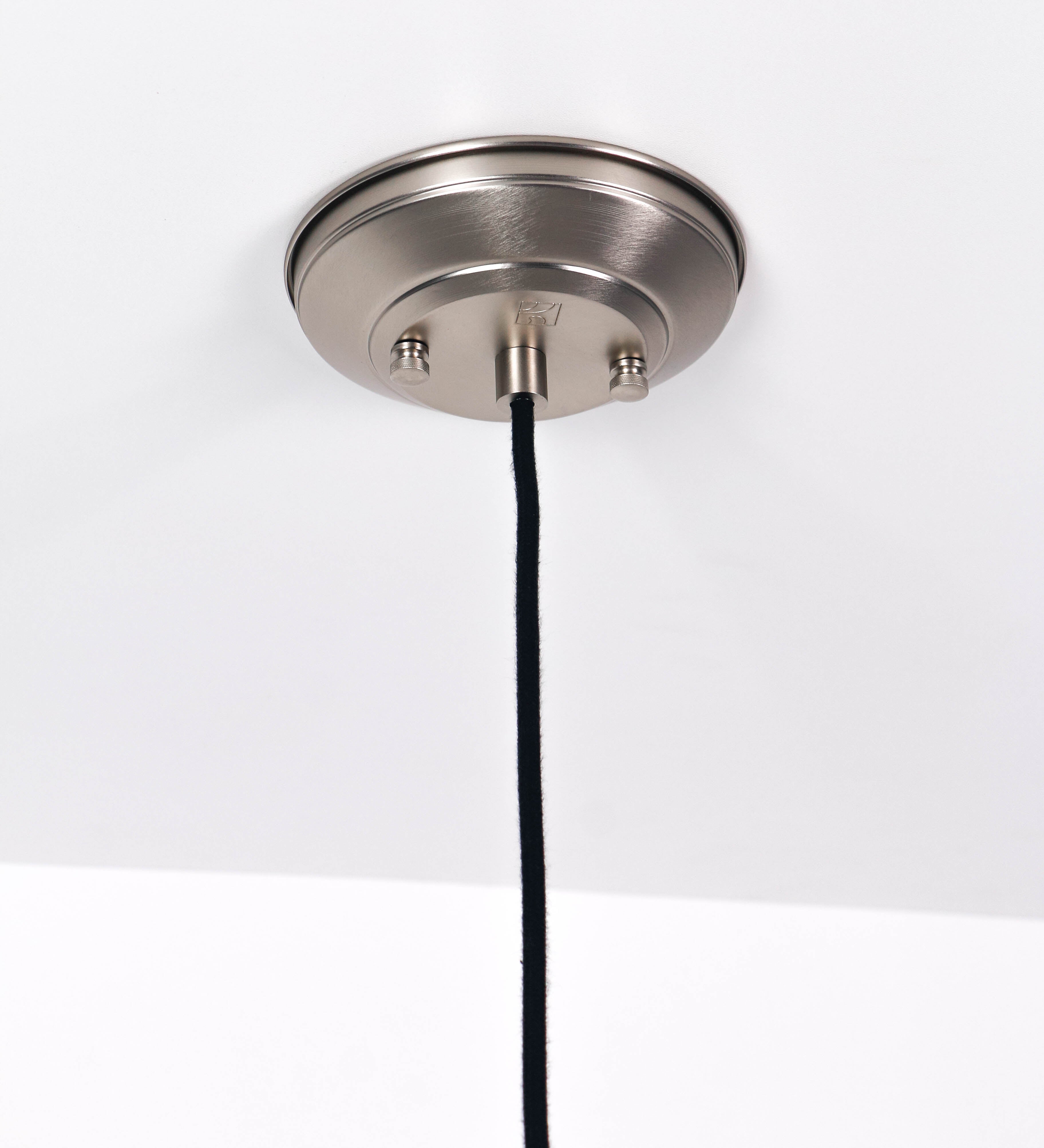 Suspensão de teto que combina um design clássico com um acabamento moderno e de estilo industrial. A suspensão, toda em níquel escovado, é fixa ao teto através de uma base circular de onde desce um cabo revestido têxtil preto, que sustenta o chapéu principal. O chapéu tem um formato cónico invertido, com uma borda ligeiramente levantada. No topo do chapéu, há um suporte circular com três pequenos braços que conecta o chapéu ao cabo.