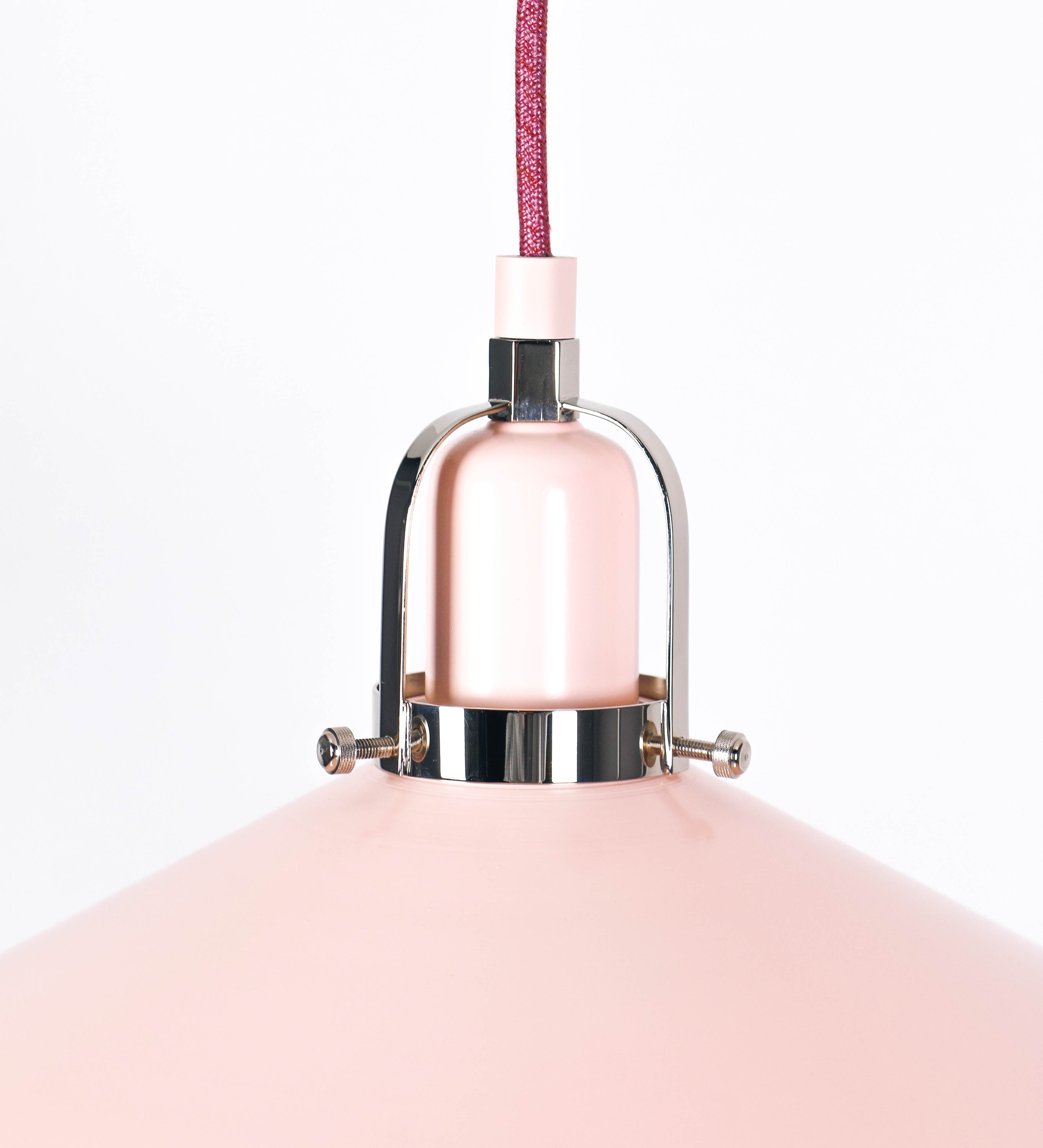 Suspensão de teto que combina um design clássico com cores modernas. A suspensão é fixa ao teto através de uma base circular rosa clara, de onde desce um cabo revestido têxtil rosa velho, que sustenta o chapéu principal. O chapéu, rosa claro, tem um formato cónico invertido, com uma borda ligeiramente levantada. O interior do chapéu é branco. No topo do chapéu, há um suporte circular em níquel polido, com três pequenos braços que conecta o chapéu ao cabo.