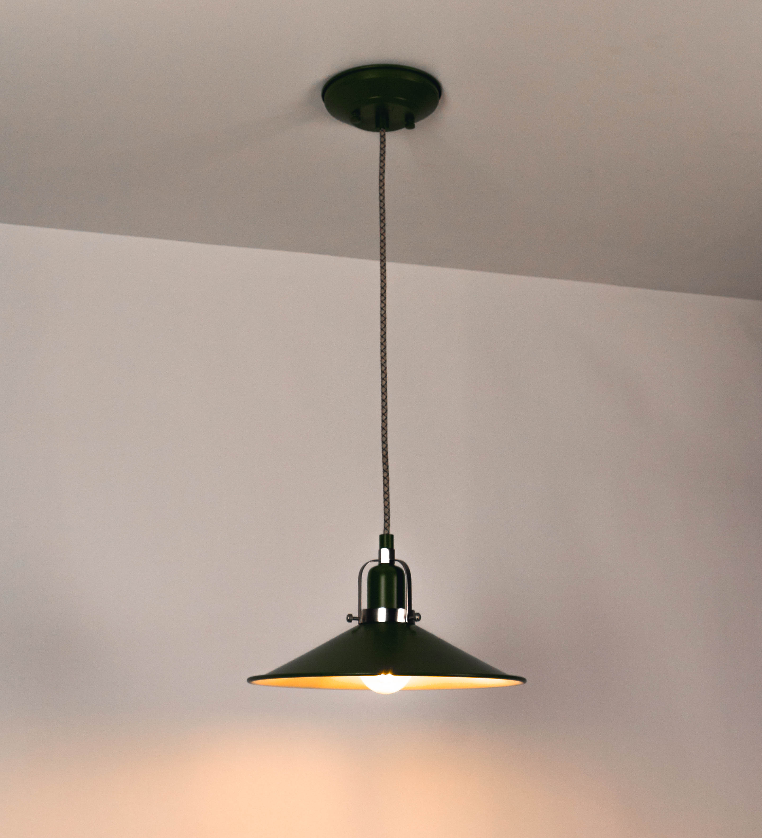 Suspensão de teto que combina um design clássico e cores modernas. A suspensão é fixa ao teto através de uma base circular verde couve-de-bruxelas, de onde desce um cabo revestido têxtil com um padrão de linhas castanhas e beges entrelaçadas, que sustenta o chapéu principal. O chapéu, verde, tem um formato cónico invertido, com uma borda ligeiramente levantada. O interior do chapéu é branco. No topo do chapéu, há um suporte circular em níquel escovado, com três pequenos braços que conecta o chapéu ao cabo.