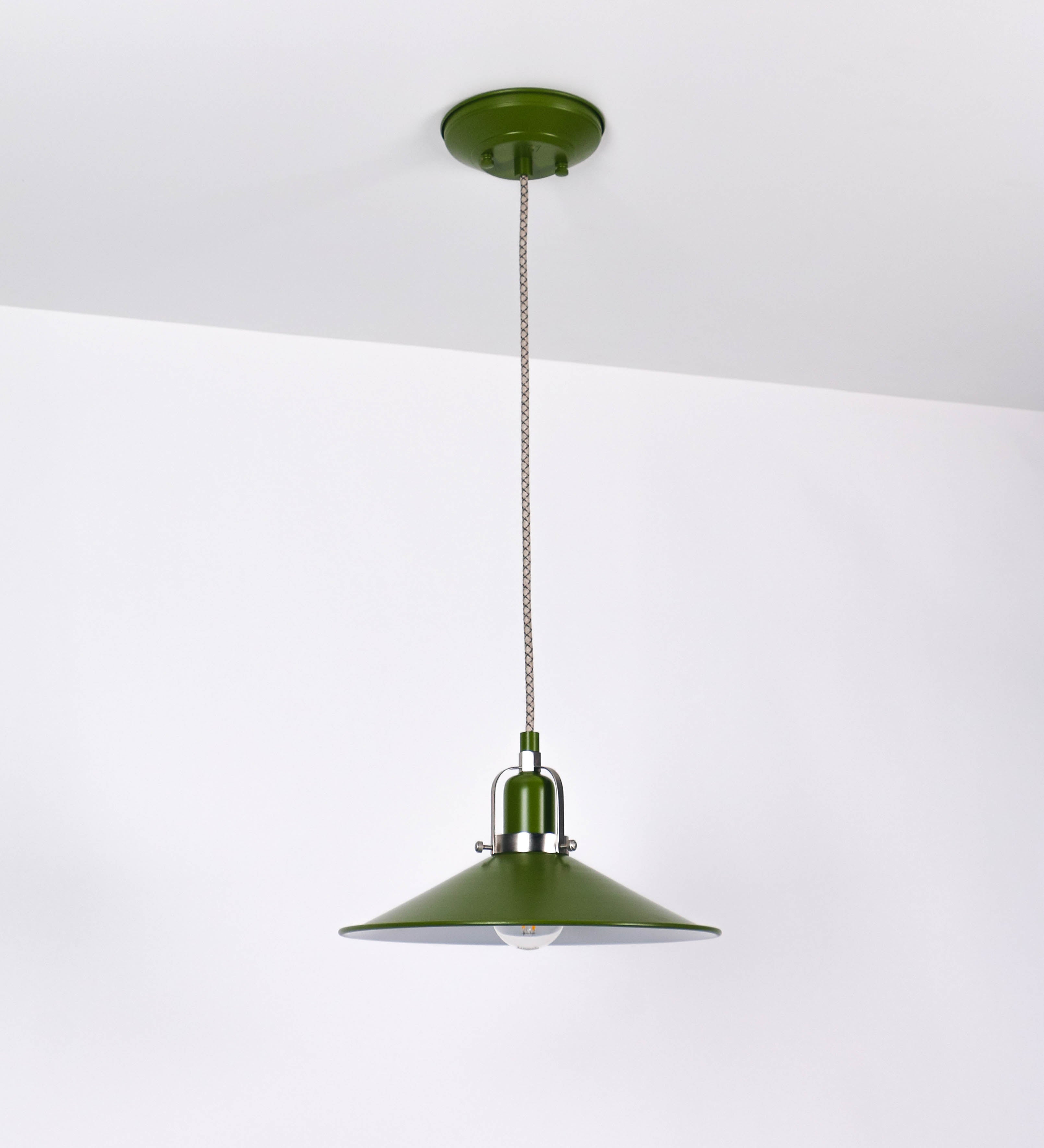 Suspensão de teto que combina um design clássico e cores modernas. A suspensão é fixa ao teto através de uma base circular verde couve-de-bruxelas, de onde desce um cabo revestido têxtil com um padrão de linhas castanhas e beges entrelaçadas, que sustenta o chapéu principal. O chapéu, verde, tem um formato cónico invertido, com uma borda ligeiramente levantada. O interior do chapéu é branco. No topo do chapéu, há um suporte circular em níquel escovado, com três pequenos braços que conecta o chapéu ao cabo.