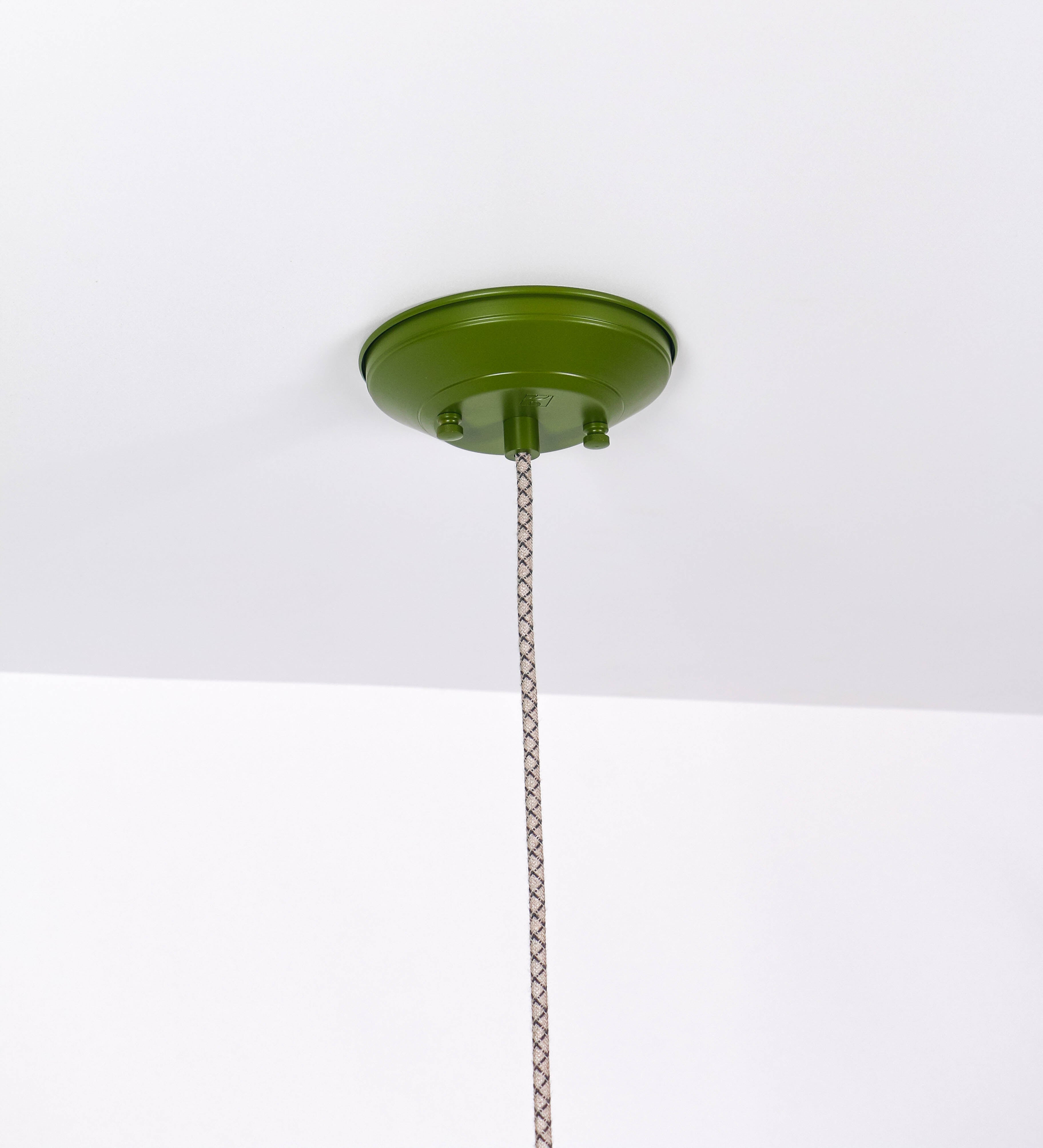 Suspensão de teto que combina um design clássico e cores modernas. A suspensão é fixa ao teto através de uma base circular verde couve-de-bruxelas, de onde desce um cabo revestido têxtil com um padrão de linhas castanhas e beges entrelaçadas, que sustenta o chapéu principal. O chapéu, verde, tem um formato cónico invertido, com uma borda ligeiramente levantada. O interior do chapéu é branco. No topo do chapéu, há um suporte circular em níquel escovado, com três pequenos braços que conecta o chapéu ao cabo.