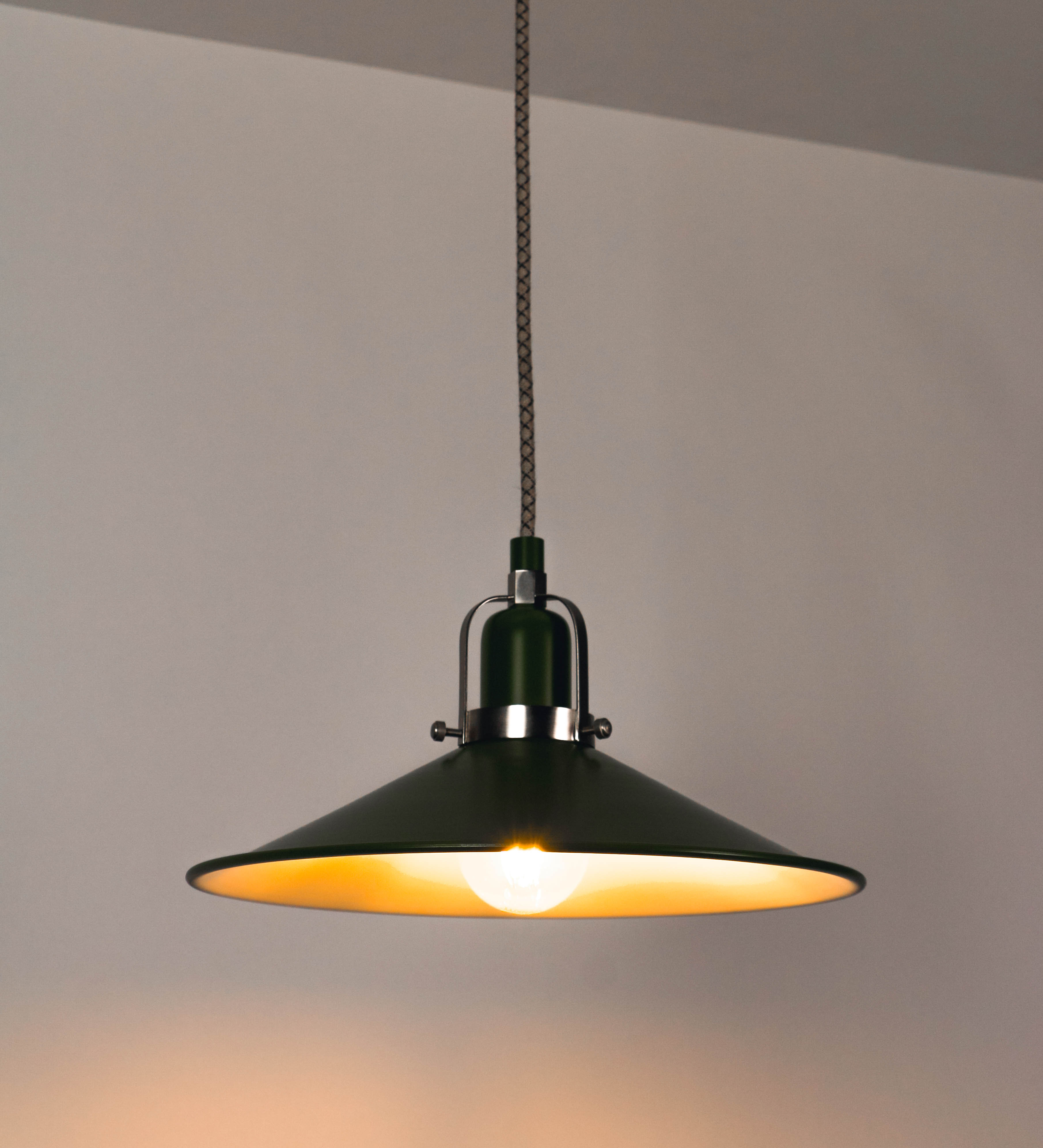 Suspensão de teto que combina um design clássico e cores modernas. A suspensão é fixa ao teto através de uma base circular verde couve-de-bruxelas, de onde desce um cabo revestido têxtil com um padrão de linhas castanhas e beges entrelaçadas, que sustenta o chapéu principal. O chapéu, verde, tem um formato cónico invertido, com uma borda ligeiramente levantada. O interior do chapéu é branco. No topo do chapéu, há um suporte circular em níquel escovado, com três pequenos braços que conecta o chapéu ao cabo.