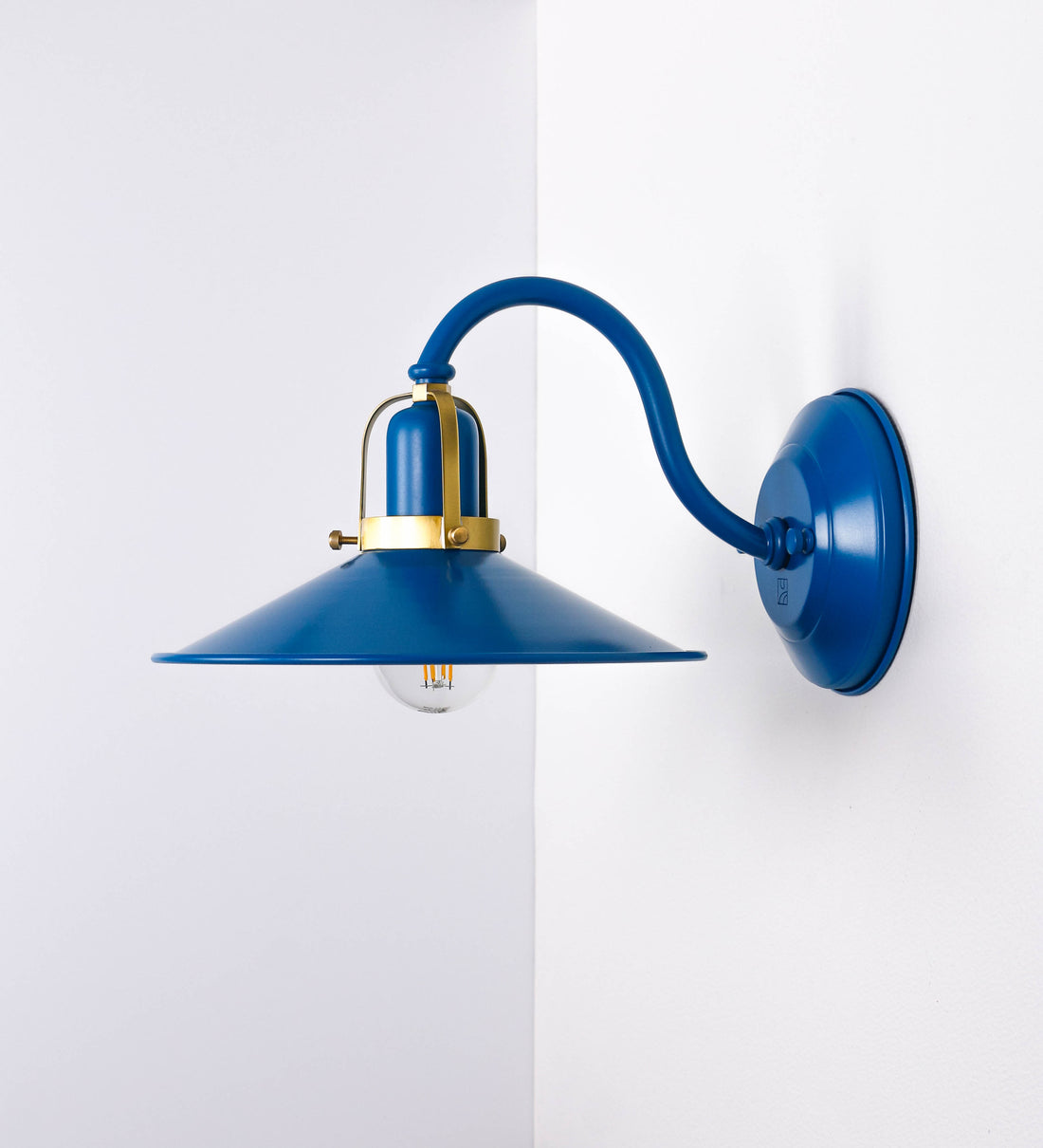 Aplique de parede com um design clássico e cores contemporâneas. O aplique é composto por uma base circular azul vintage, de onde se estende um braço curvo, que forma um arco gracioso e sustenta o chapéu do candeeiro, azul vintage também. O chapéu tem um formato cónico invertido, com uma borda ligeiramente levantada, que direciona a luz para baixo. O interior do chapéu é branco. No topo do chapéu, há um suporte circular em latão escovado, com três braços que fixa o chapéu ao braço curvo.