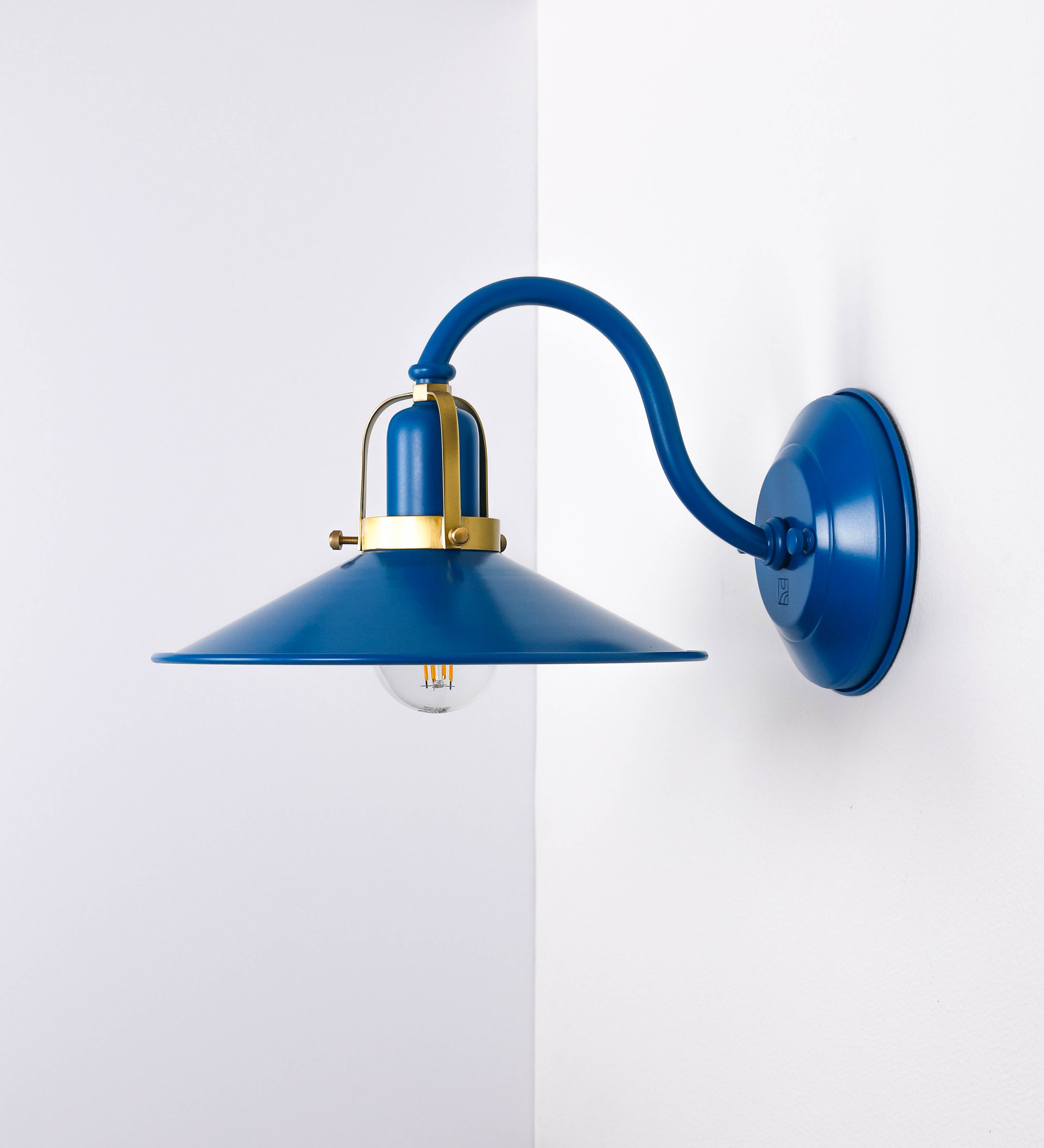 Aplique de parede com um design clássico e cores contemporâneas. O aplique é composto por uma base circular azul vintage, de onde se estende um braço curvo, que forma um arco gracioso e sustenta o chapéu do candeeiro, azul vintage também. O chapéu tem um formato cónico invertido, com uma borda ligeiramente levantada, que direciona a luz para baixo. O interior do chapéu é branco. No topo do chapéu, há um suporte circular em latão escovado, com três braços que fixa o chapéu ao braço curvo.