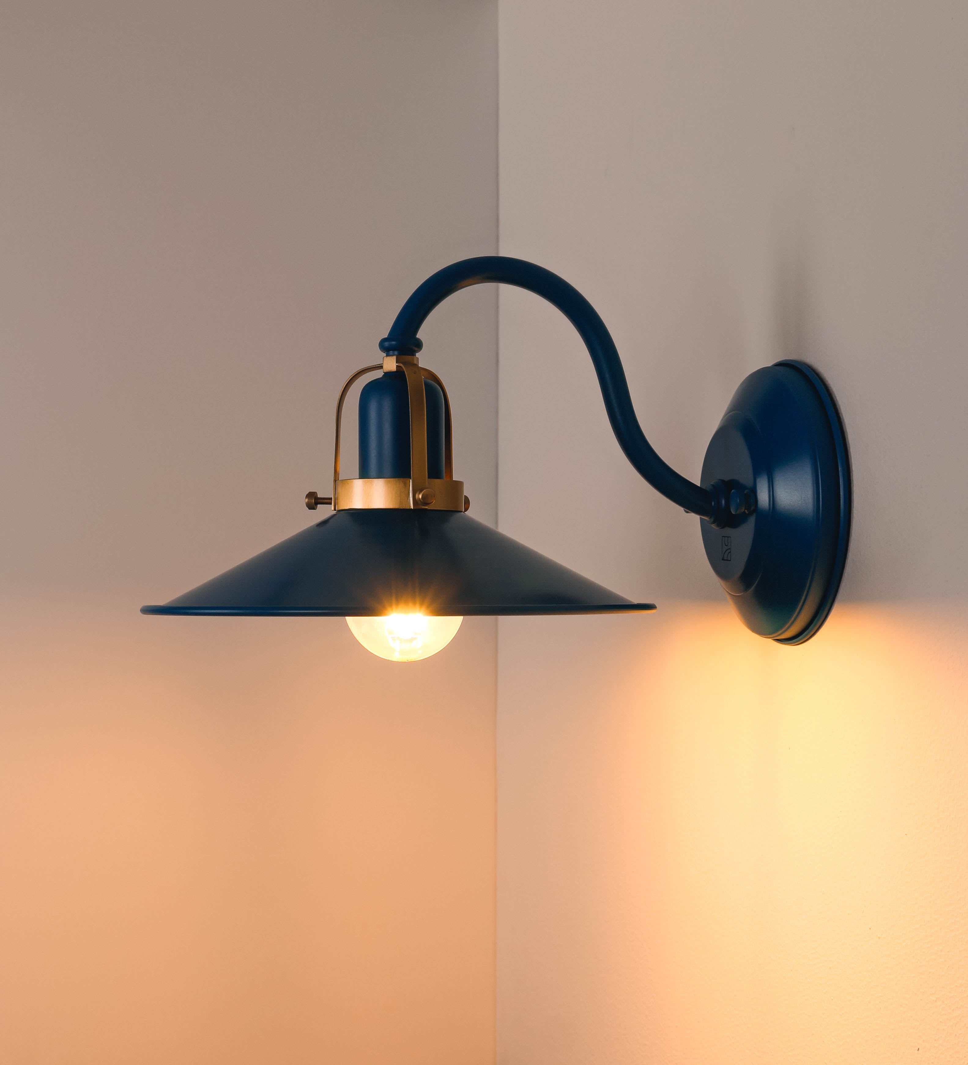 Aplique de parede com um design clássico e cores contemporâneas. O aplique é composto por uma base circular azul vintage, de onde se estende um braço curvo, que forma um arco gracioso e sustenta o chapéu do candeeiro, azul vintage também. O chapéu tem um formato cónico invertido, com uma borda ligeiramente levantada, que direciona a luz para baixo. O interior do chapéu é branco. No topo do chapéu, há um suporte circular em latão escovado, com três braços que fixa o chapéu ao braço curvo.