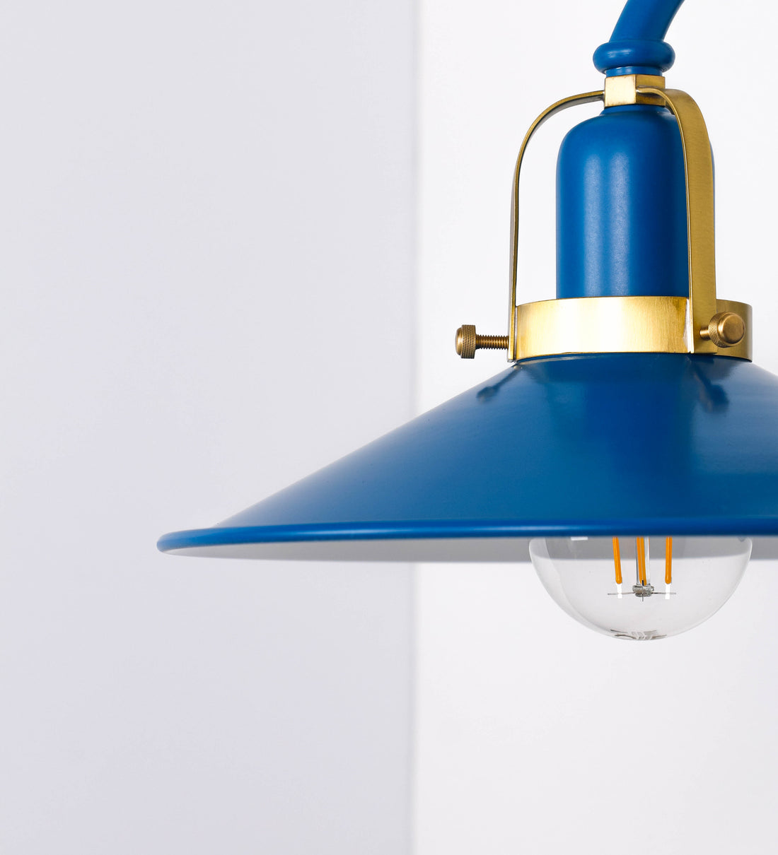 Aplique de parede com um design clássico e cores contemporâneas. O aplique é composto por uma base circular azul vintage, de onde se estende um braço curvo, que forma um arco gracioso e sustenta o chapéu do candeeiro, azul vintage também. O chapéu tem um formato cónico invertido, com uma borda ligeiramente levantada, que direciona a luz para baixo. O interior do chapéu é branco. No topo do chapéu, há um suporte circular em latão escovado, com três braços que fixa o chapéu ao braço curvo.