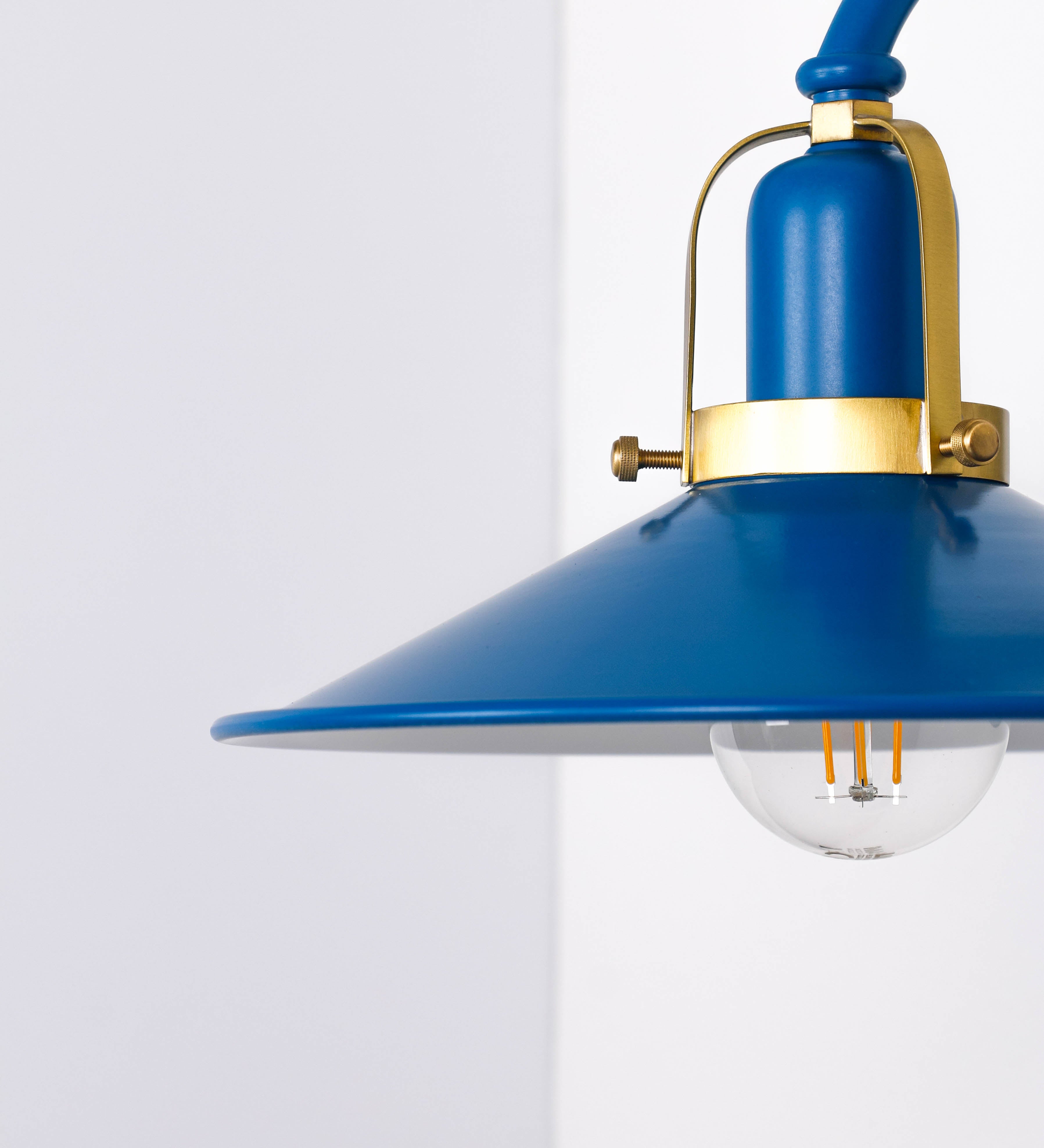Aplique de parede com um design clássico e cores contemporâneas. O aplique é composto por uma base circular azul vintage, de onde se estende um braço curvo, que forma um arco gracioso e sustenta o chapéu do candeeiro, azul vintage também. O chapéu tem um formato cónico invertido, com uma borda ligeiramente levantada, que direciona a luz para baixo. O interior do chapéu é branco. No topo do chapéu, há um suporte circular em latão escovado, com três braços que fixa o chapéu ao braço curvo.
