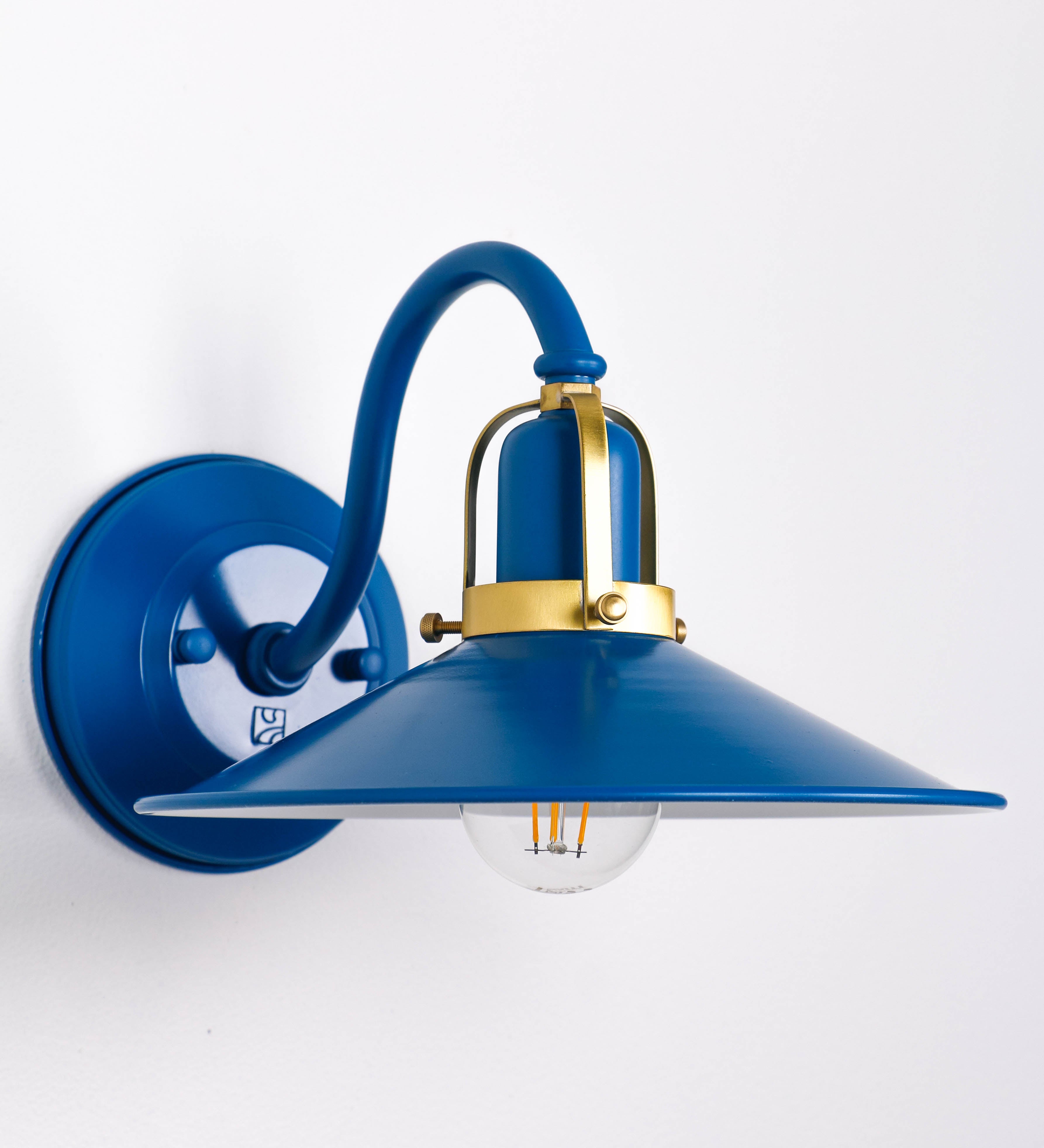 Aplique de parede com um design clássico e cores contemporâneas. O aplique é composto por uma base circular azul vintage, de onde se estende um braço curvo, que forma um arco gracioso e sustenta o chapéu do candeeiro, azul vintage também. O chapéu tem um formato cónico invertido, com uma borda ligeiramente levantada, que direciona a luz para baixo. O interior do chapéu é branco. No topo do chapéu, há um suporte circular em latão escovado, com três braços que fixa o chapéu ao braço curvo.