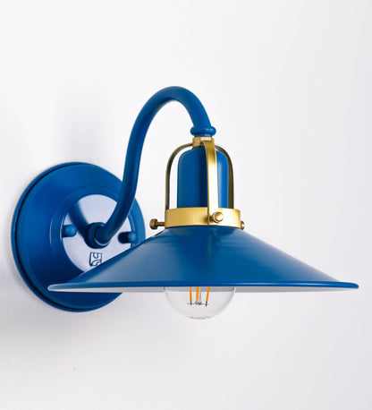 Aplique de parede com um design clássico e cores contemporâneas. O aplique é composto por uma base circular azul vintage, de onde se estende um braço curvo, que forma um arco gracioso e sustenta o chapéu do candeeiro, azul vintage também. O chapéu tem um formato cónico invertido, com uma borda ligeiramente levantada, que direciona a luz para baixo. O interior do chapéu é branco. No topo do chapéu, há um suporte circular em latão escovado, com três braços que fixa o chapéu ao braço curvo.