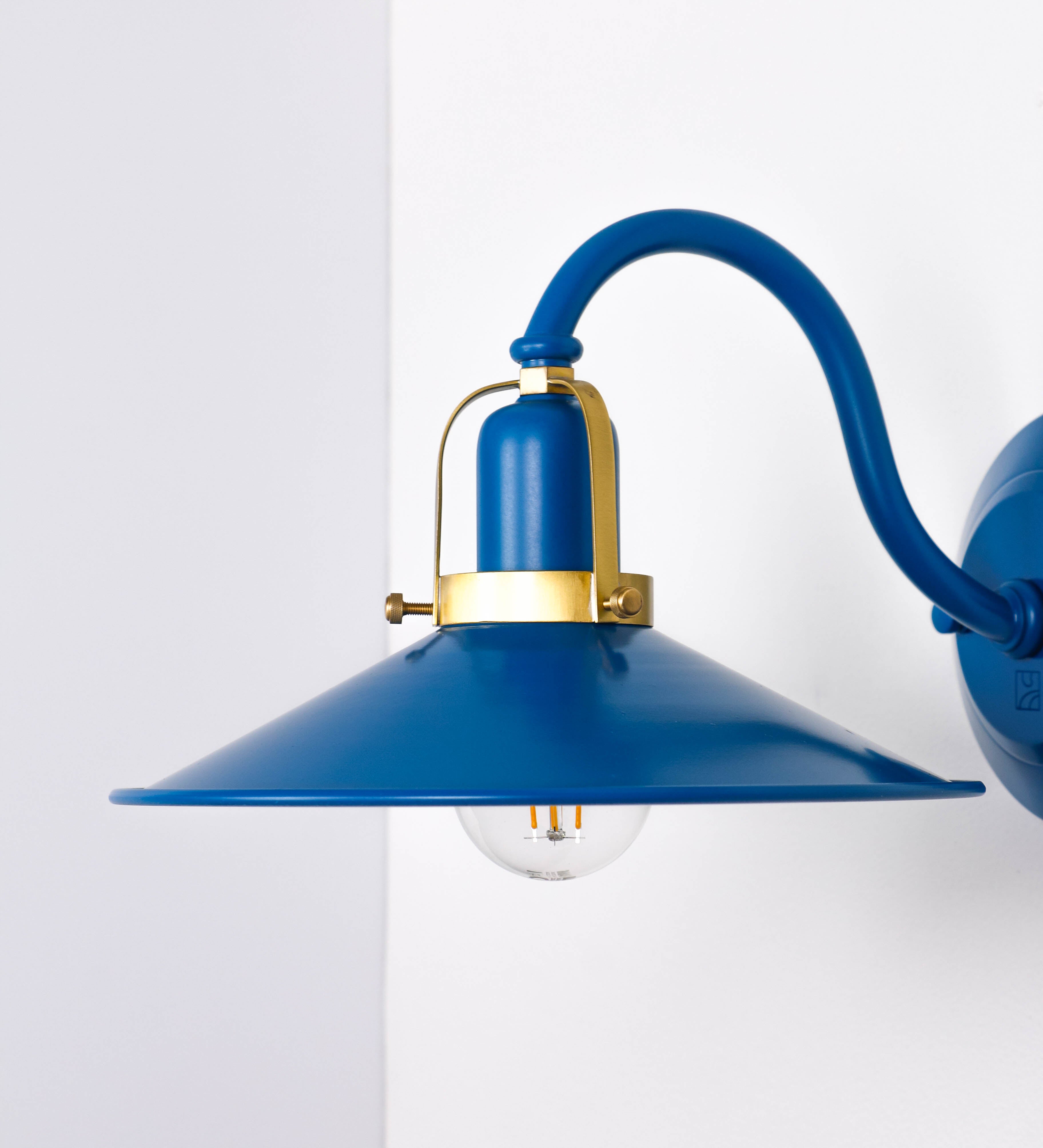 Aplique de parede com um design clássico e cores contemporâneas. O aplique é composto por uma base circular azul vintage, de onde se estende um braço curvo, que forma um arco gracioso e sustenta o chapéu do candeeiro, azul vintage também. O chapéu tem um formato cónico invertido, com uma borda ligeiramente levantada, que direciona a luz para baixo. O interior do chapéu é branco. No topo do chapéu, há um suporte circular em latão escovado, com três braços que fixa o chapéu ao braço curvo.