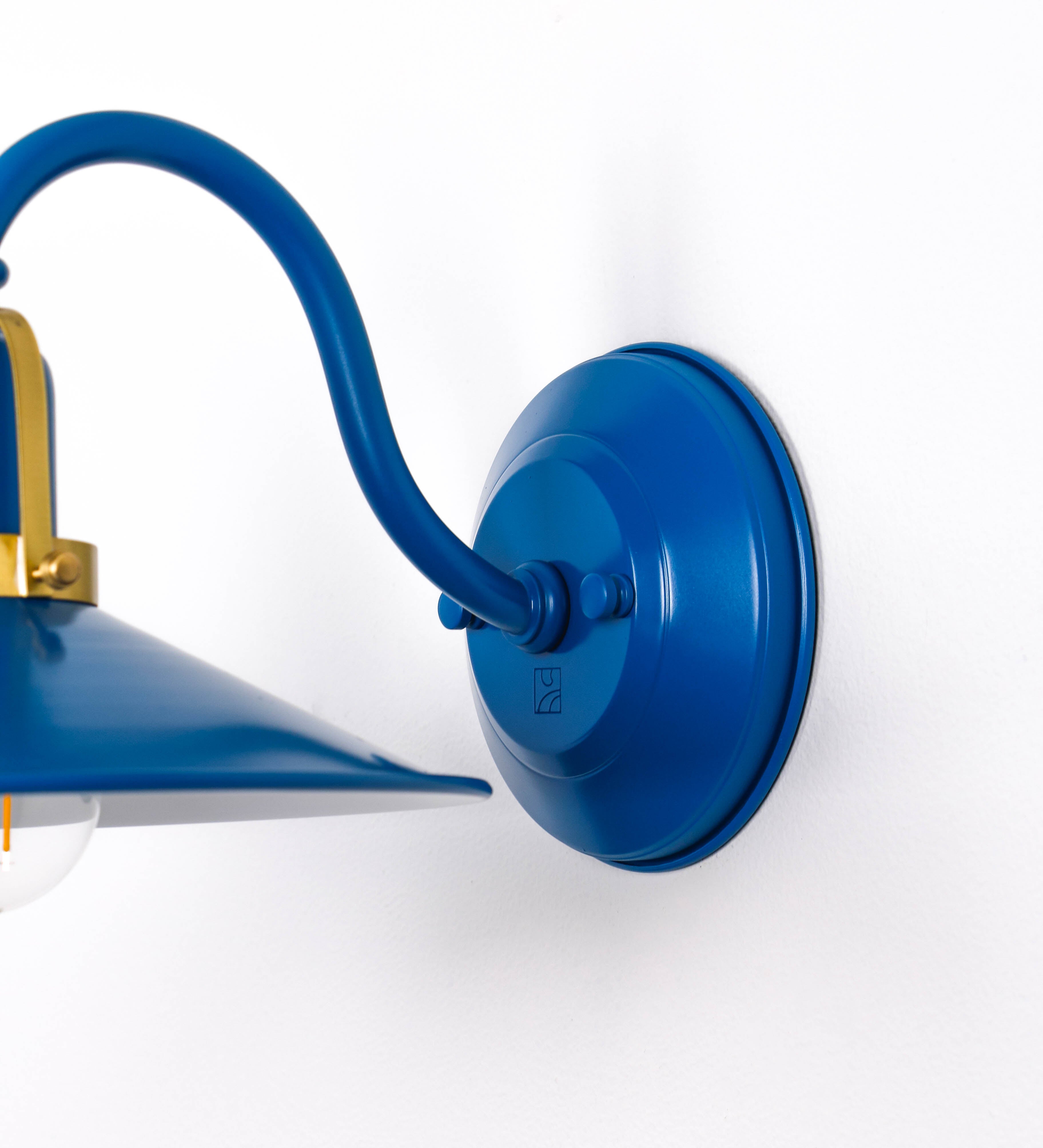 Aplique de parede com um design clássico e cores contemporâneas. O aplique é composto por uma base circular azul vintage, de onde se estende um braço curvo, que forma um arco gracioso e sustenta o chapéu do candeeiro, azul vintage também. O chapéu tem um formato cónico invertido, com uma borda ligeiramente levantada, que direciona a luz para baixo. O interior do chapéu é branco. No topo do chapéu, há um suporte circular em latão escovado, com três braços que fixa o chapéu ao braço curvo.