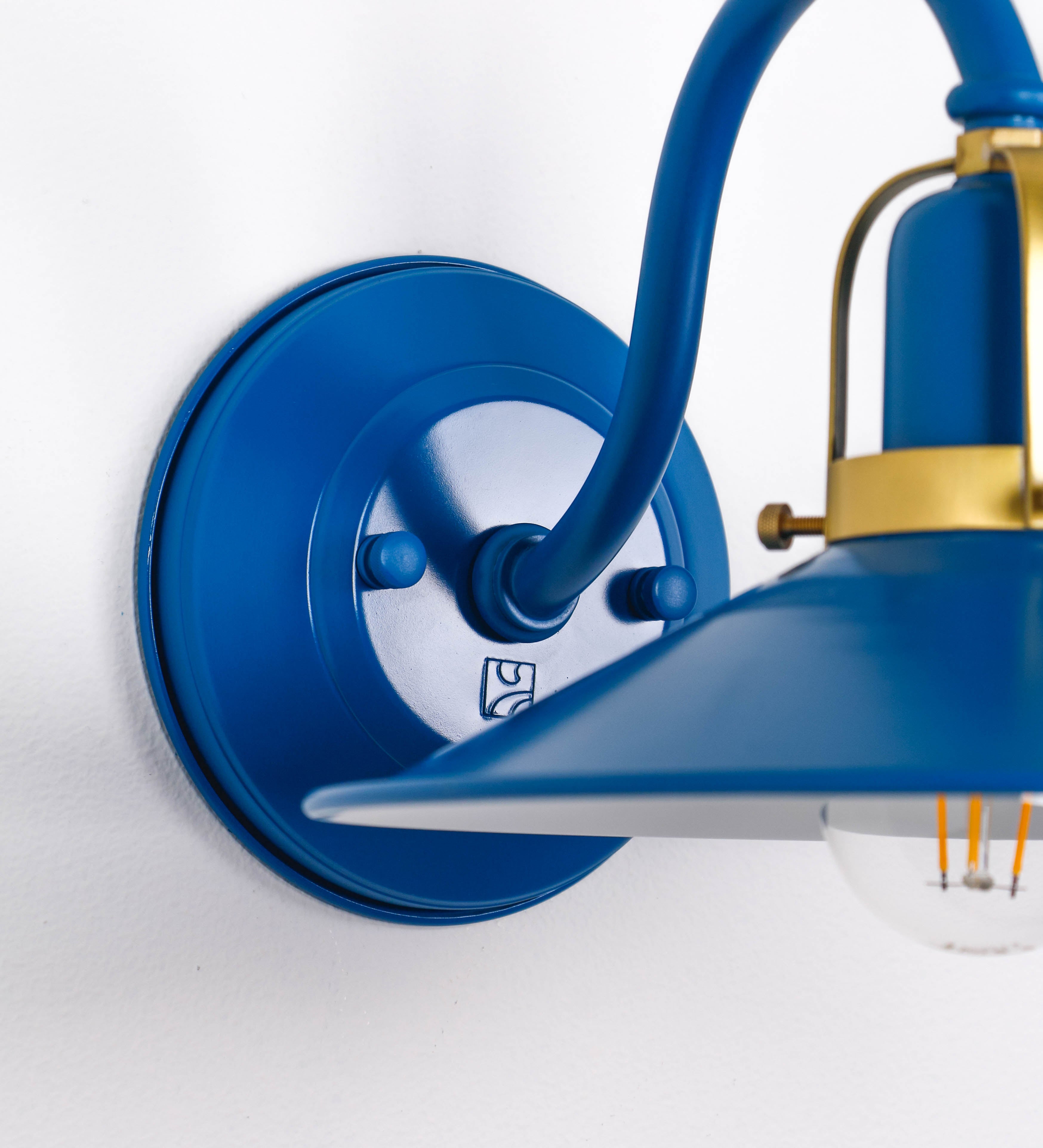 Aplique de parede com um design clássico e cores contemporâneas. O aplique é composto por uma base circular azul vintage, de onde se estende um braço curvo, que forma um arco gracioso e sustenta o chapéu do candeeiro, azul vintage também. O chapéu tem um formato cónico invertido, com uma borda ligeiramente levantada, que direciona a luz para baixo. O interior do chapéu é branco. No topo do chapéu, há um suporte circular em latão escovado, com três braços que fixa o chapéu ao braço curvo.