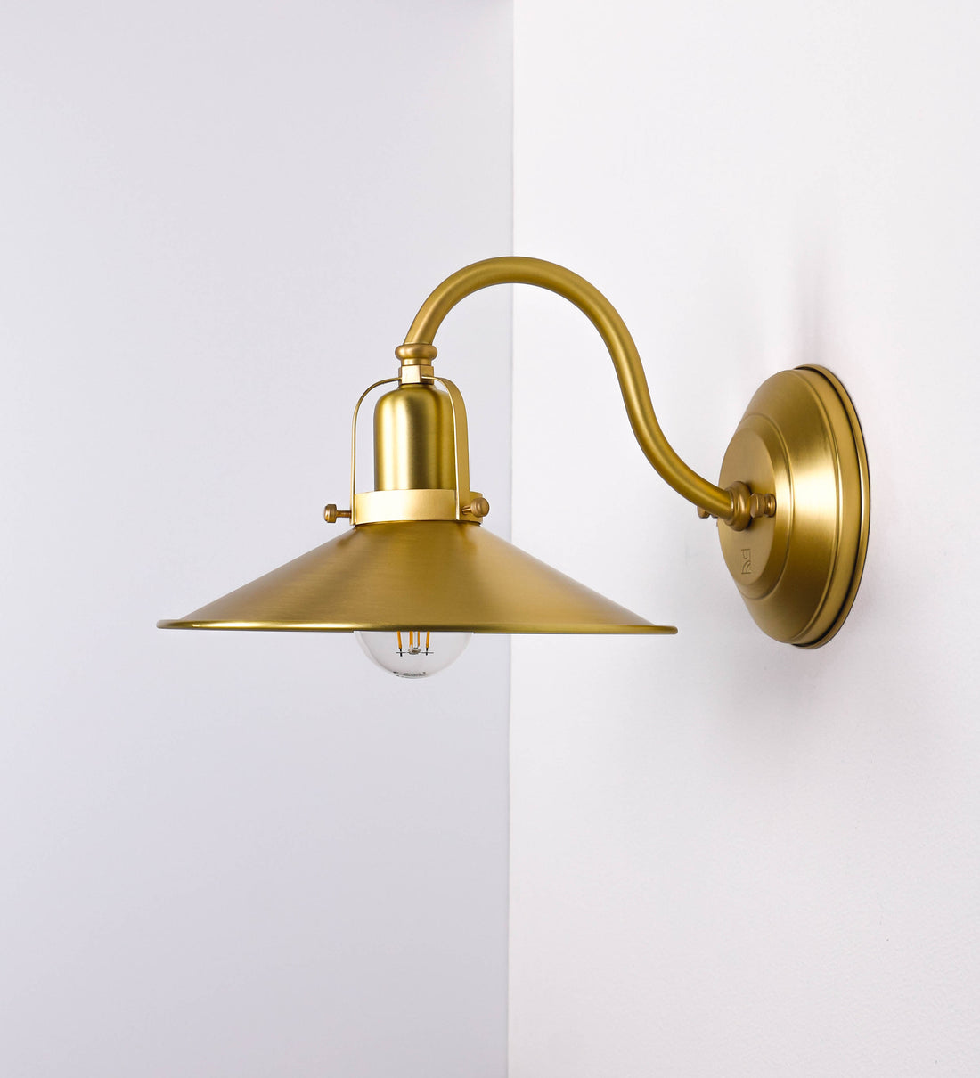 Aplique de parede todo em latão escovado, com um design clássico e cores contemporâneas. O aplique é composto por uma base circular, de onde se estende um braço curvo, que forma um arco gracioso e sustenta o chapéu do candeeiro. O chapéu tem um formato cónico invertido, com uma borda ligeiramente levantada, que direciona a luz para baixo. No topo do chapéu, há um suporte circular com três braços que fixa o chapéu ao braço curvo.
