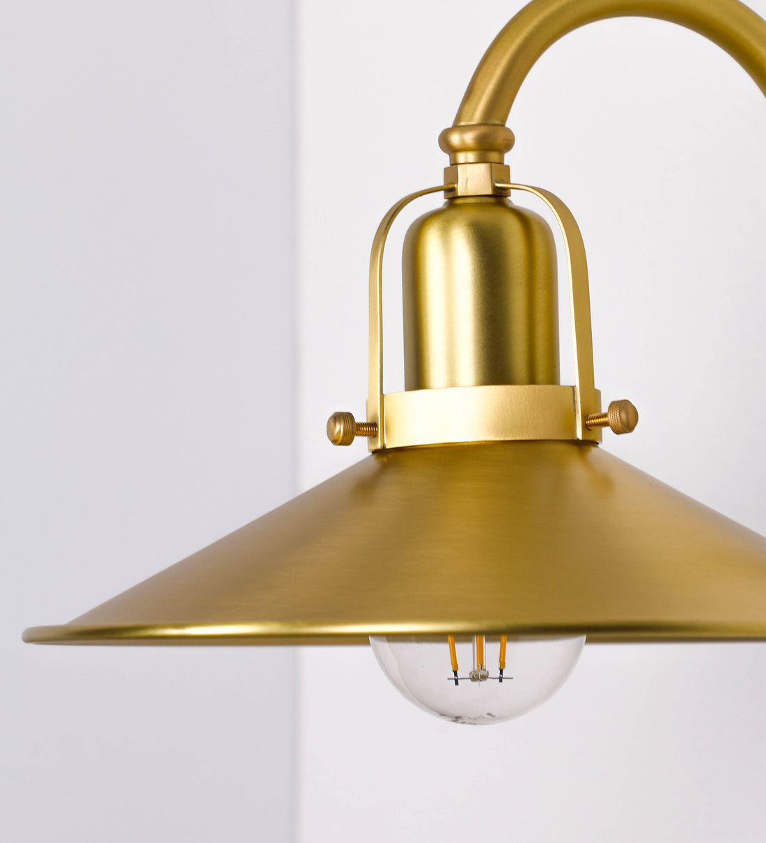 Aplique de parede todo em latão escovado, com um design clássico e cores contemporâneas. O aplique é composto por uma base circular, de onde se estende um braço curvo, que forma um arco gracioso e sustenta o chapéu do candeeiro. O chapéu tem um formato cónico invertido, com uma borda ligeiramente levantada, que direciona a luz para baixo. No topo do chapéu, há um suporte circular com três braços que fixa o chapéu ao braço curvo.