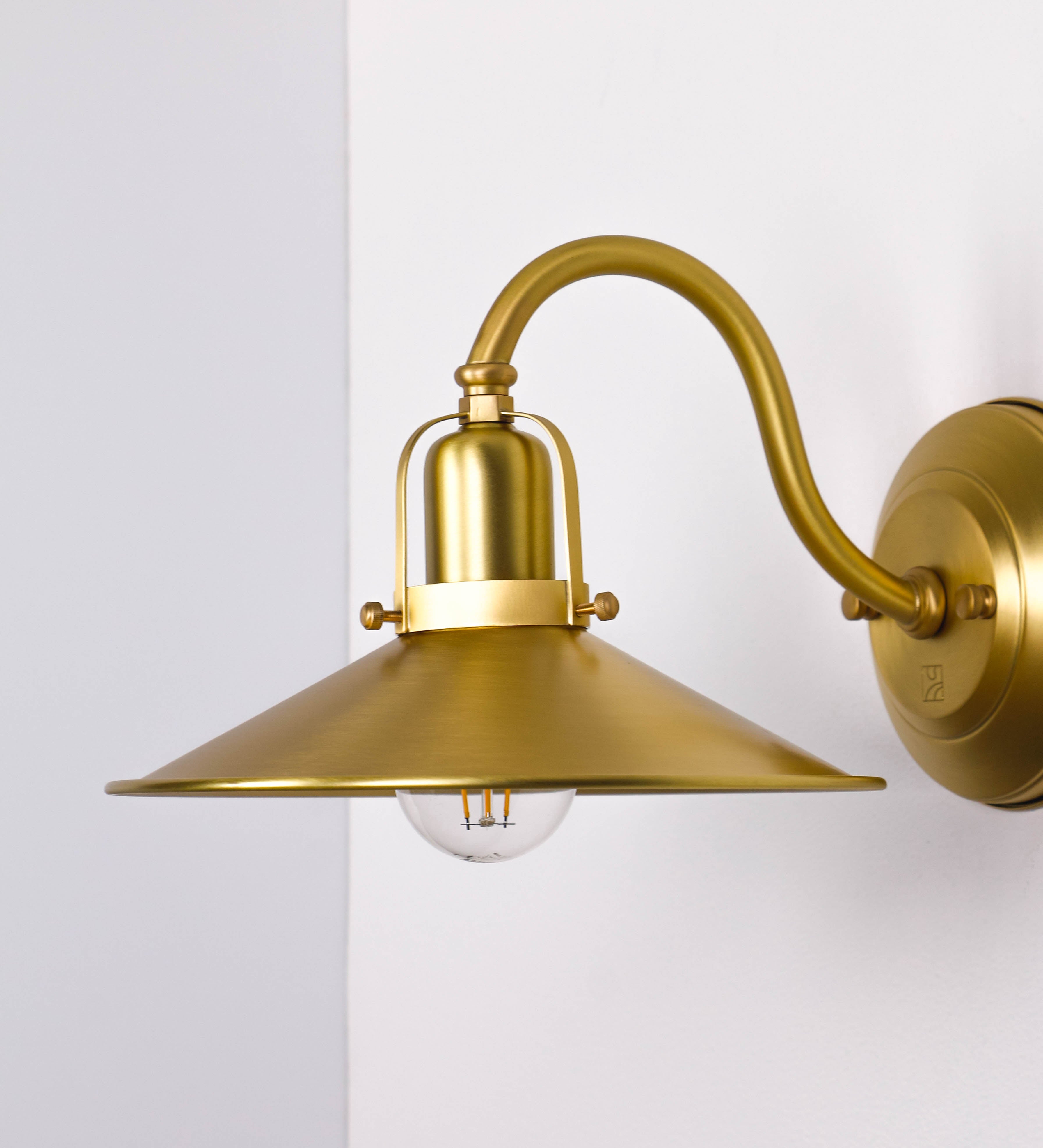 Aplique de parede todo em latão escovado, com um design clássico e cores contemporâneas. O aplique é composto por uma base circular, de onde se estende um braço curvo, que forma um arco gracioso e sustenta o chapéu do candeeiro. O chapéu tem um formato cónico invertido, com uma borda ligeiramente levantada, que direciona a luz para baixo. No topo do chapéu, há um suporte circular com três braços que fixa o chapéu ao braço curvo.