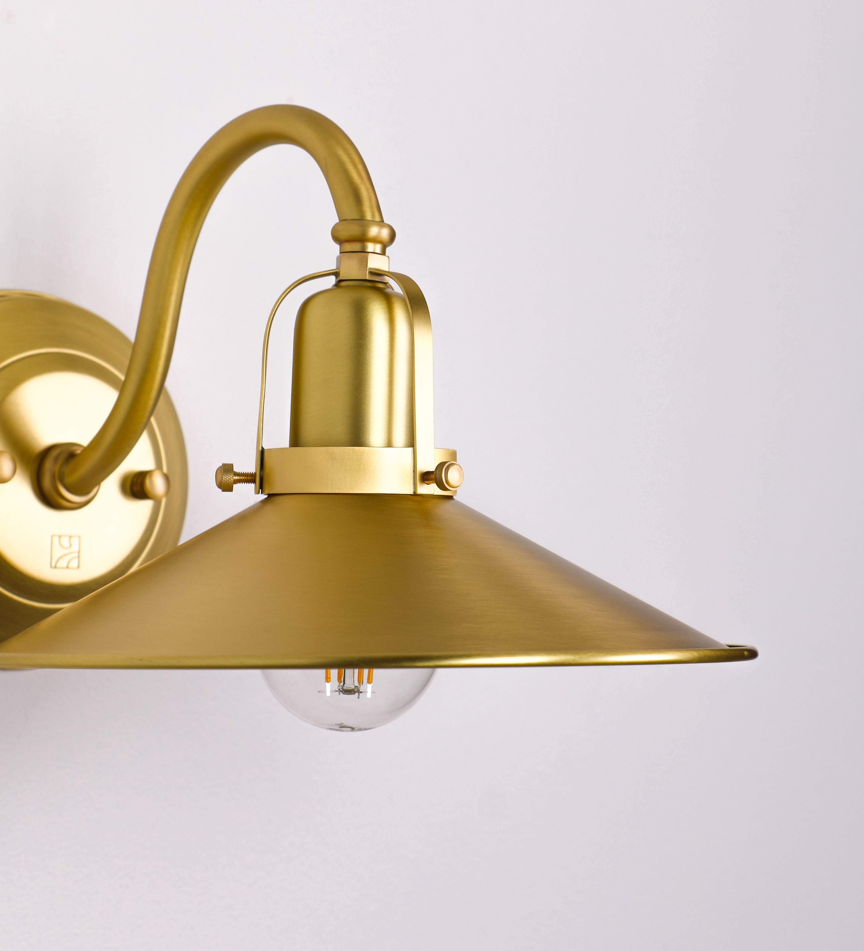 Aplique de parede todo em latão escovado, com um design clássico e cores contemporâneas. O aplique é composto por uma base circular, de onde se estende um braço curvo, que forma um arco gracioso e sustenta o chapéu do candeeiro. O chapéu tem um formato cónico invertido, com uma borda ligeiramente levantada, que direciona a luz para baixo. No topo do chapéu, há um suporte circular com três braços que fixa o chapéu ao braço curvo.