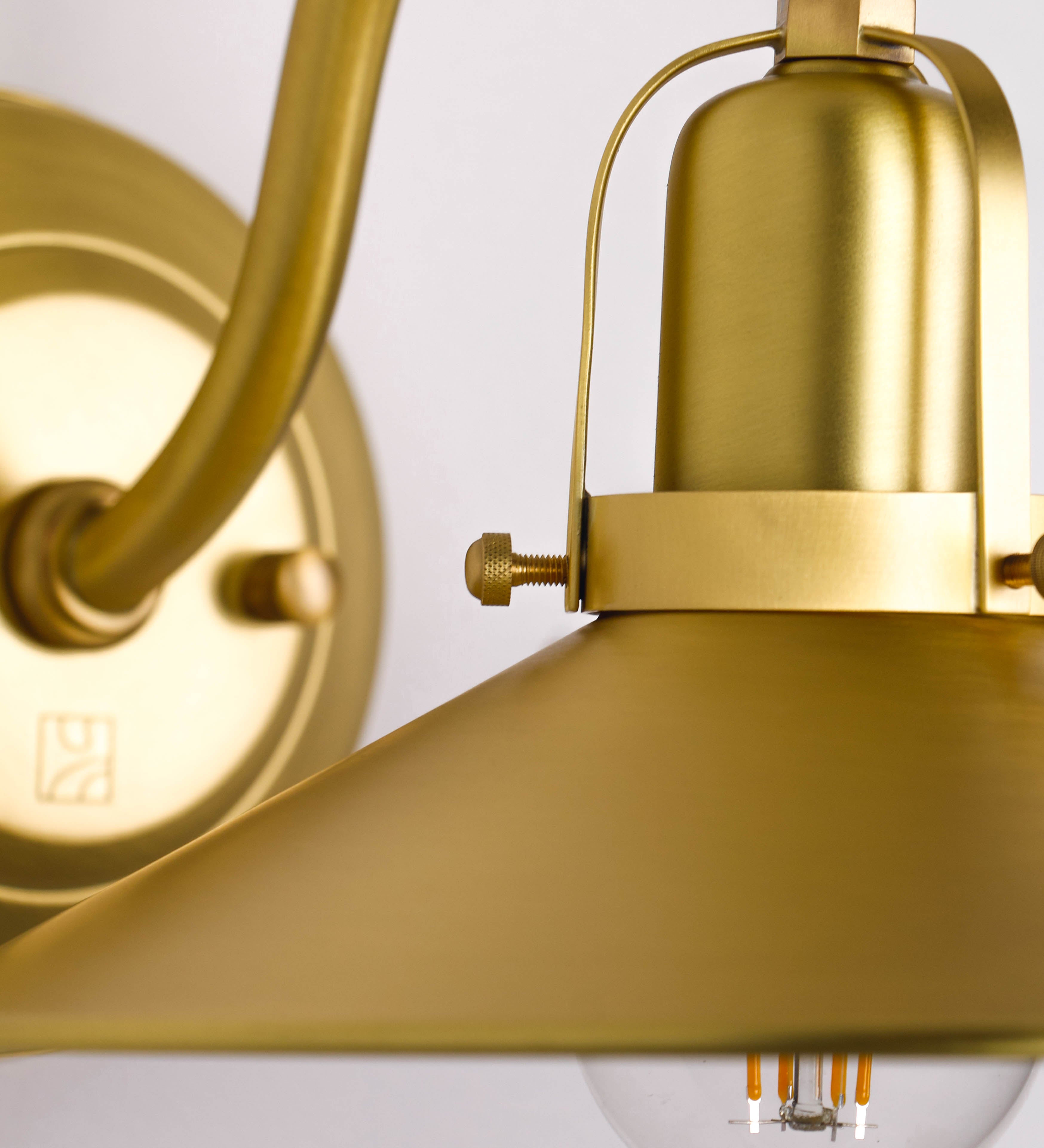 Aplique de parede todo em latão escovado, com um design clássico e cores contemporâneas. O aplique é composto por uma base circular, de onde se estende um braço curvo, que forma um arco gracioso e sustenta o chapéu do candeeiro. O chapéu tem um formato cónico invertido, com uma borda ligeiramente levantada, que direciona a luz para baixo. No topo do chapéu, há um suporte circular com três braços que fixa o chapéu ao braço curvo.