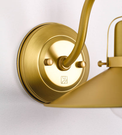 Aplique de parede todo em latão escovado, com um design clássico e cores contemporâneas. O aplique é composto por uma base circular, de onde se estende um braço curvo, que forma um arco gracioso e sustenta o chapéu do candeeiro. O chapéu tem um formato cónico invertido, com uma borda ligeiramente levantada, que direciona a luz para baixo. No topo do chapéu, há um suporte circular com três braços que fixa o chapéu ao braço curvo.