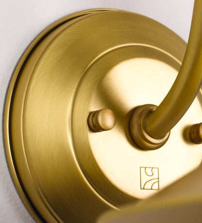 Aplique de parede todo em latão escovado, com um design clássico e cores contemporâneas. O aplique é composto por uma base circular, de onde se estende um braço curvo, que forma um arco gracioso e sustenta o chapéu do candeeiro. O chapéu tem um formato cónico invertido, com uma borda ligeiramente levantada, que direciona a luz para baixo. No topo do chapéu, há um suporte circular com três braços que fixa o chapéu ao braço curvo.