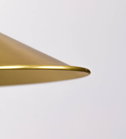 Aplique de parede todo em latão escovado, com um design clássico e cores contemporâneas. O aplique é composto por uma base circular, de onde se estende um braço curvo, que forma um arco gracioso e sustenta o chapéu do candeeiro. O chapéu tem um formato cónico invertido, com uma borda ligeiramente levantada, que direciona a luz para baixo. No topo do chapéu, há um suporte circular com três braços que fixa o chapéu ao braço curvo.