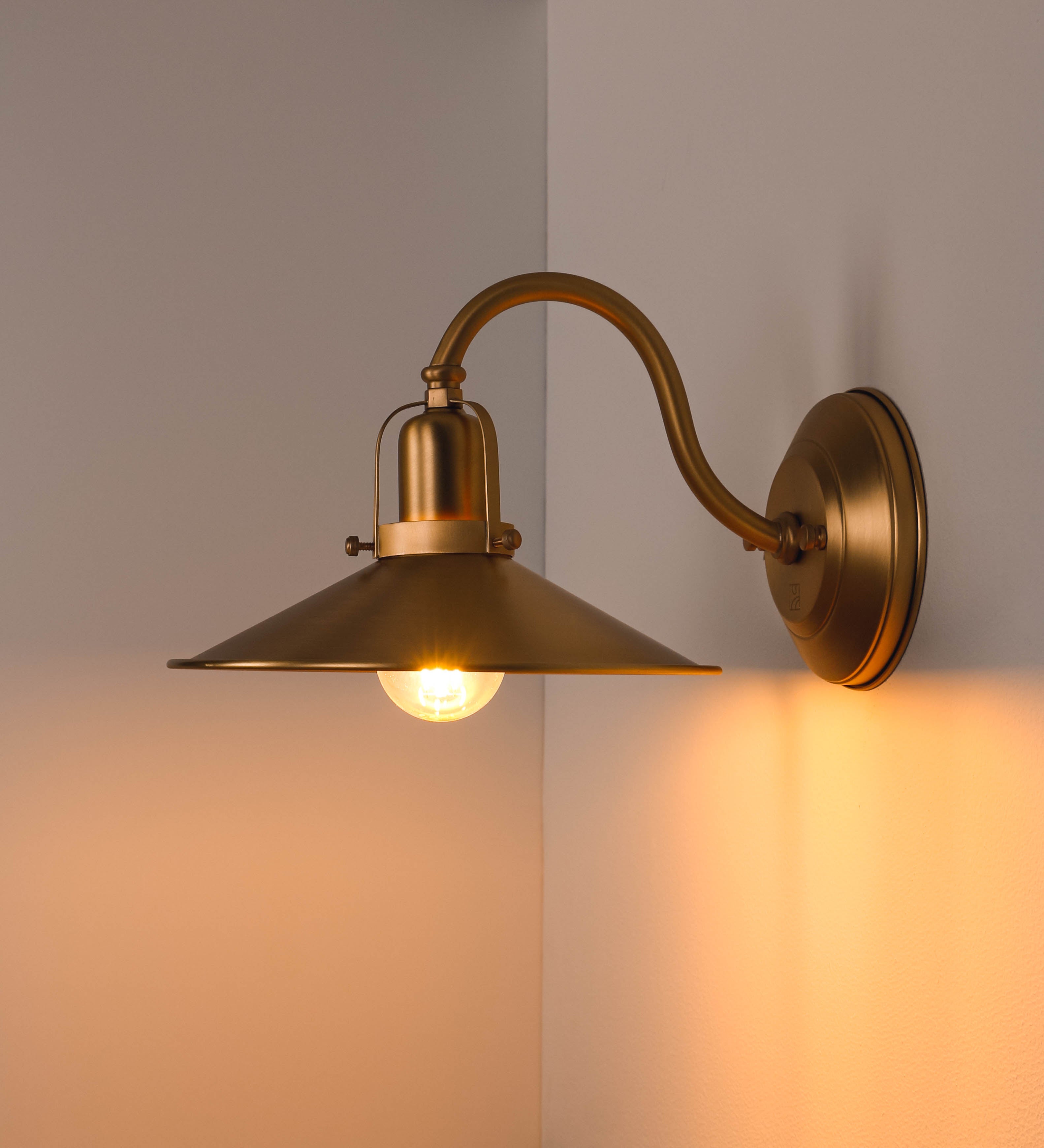 Aplique de parede todo em latão escovado, com um design clássico e cores contemporâneas. O aplique é composto por uma base circular, de onde se estende um braço curvo, que forma um arco gracioso e sustenta o chapéu do candeeiro. O chapéu tem um formato cónico invertido, com uma borda ligeiramente levantada, que direciona a luz para baixo. No topo do chapéu, há um suporte circular com três braços que fixa o chapéu ao braço curvo.