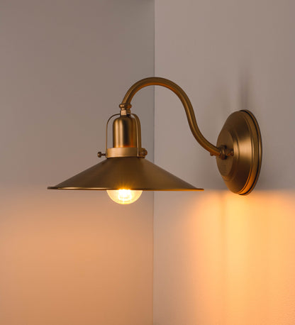 Aplique de parede todo em latão escovado, com um design clássico e cores contemporâneas. O aplique é composto por uma base circular, de onde se estende um braço curvo, que forma um arco gracioso e sustenta o chapéu do candeeiro. O chapéu tem um formato cónico invertido, com uma borda ligeiramente levantada, que direciona a luz para baixo. No topo do chapéu, há um suporte circular com três braços que fixa o chapéu ao braço curvo.
