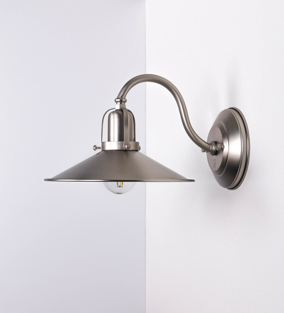 Aplique de parede todo em níquel escovado, com um design clássico e cores contemporâneas. O aplique é composto por uma base circular, de onde se estende um braço curvo, que forma um arco gracioso e sustenta o chapéu do candeeiro. O chapéu tem um formato cónico invertido, com uma borda ligeiramente levantada, que direciona a luz para baixo. No topo do chapéu, há um suporte circular com três braços que fixa o chapéu ao braço curvo.
