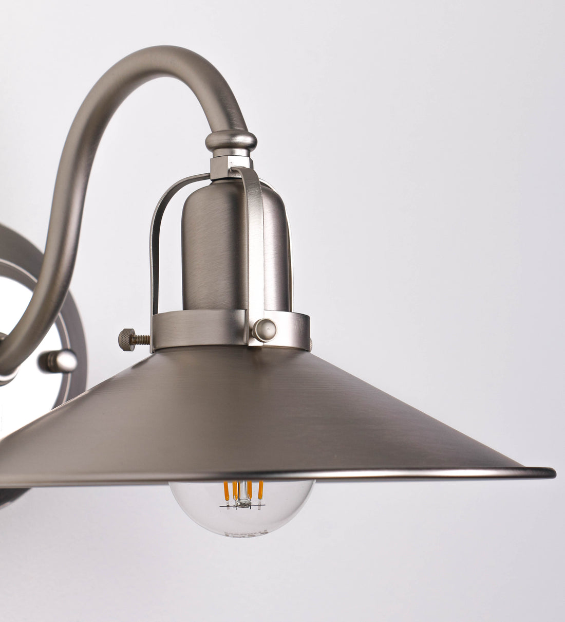 Aplique de parede todo em níquel escovado, com um design clássico e cores contemporâneas. O aplique é composto por uma base circular, de onde se estende um braço curvo, que forma um arco gracioso e sustenta o chapéu do candeeiro. O chapéu tem um formato cónico invertido, com uma borda ligeiramente levantada, que direciona a luz para baixo. No topo do chapéu, há um suporte circular com três braços que fixa o chapéu ao braço curvo.