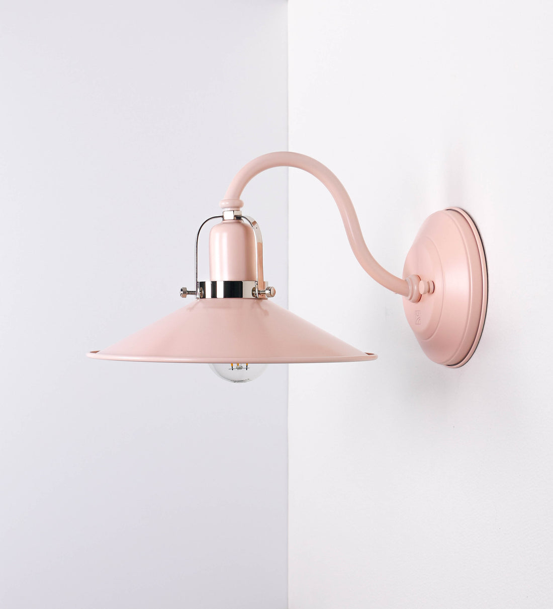 Aplique de parede com um design clássico e cores contemporâneas. O aplique é composto por uma base circular rosa clara, de onde se estende um braço curvo, que forma um arco gracioso e sustenta o chapéu do candeeiro, rosa claro também. O chapéu tem um formato cónico invertido, com uma borda ligeiramente levantada, que direciona a luz para baixo. O interior do chapéu é branco. No topo do chapéu, há um suporte circular em níquel polido, com três braços que fixa o chapéu ao braço curvo.