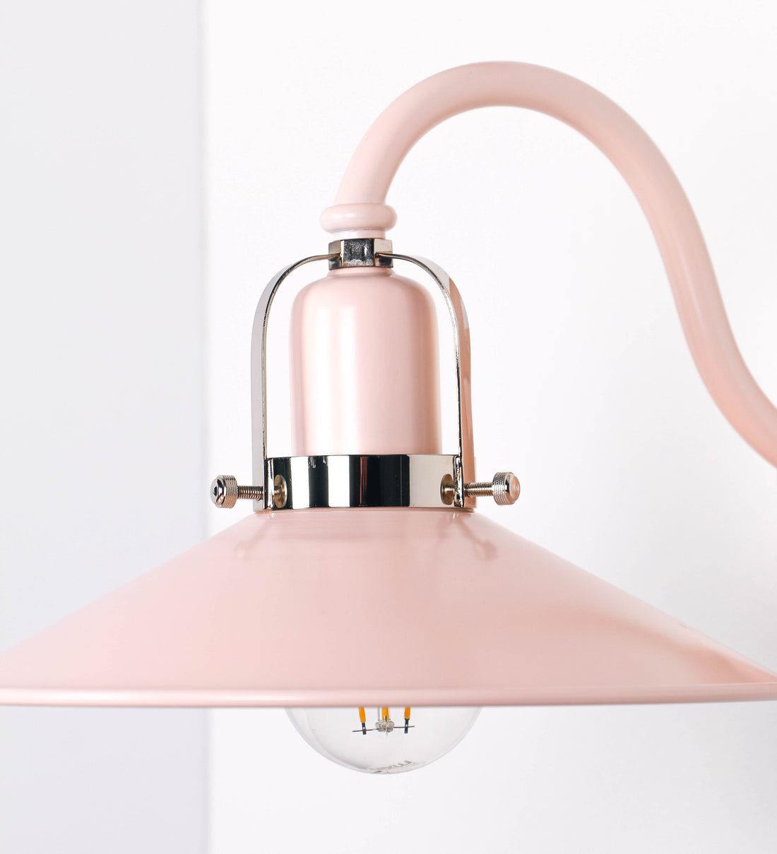 Aplique de parede com um design clássico e cores contemporâneas. O aplique é composto por uma base circular rosa clara, de onde se estende um braço curvo, que forma um arco gracioso e sustenta o chapéu do candeeiro, rosa claro também. O chapéu tem um formato cónico invertido, com uma borda ligeiramente levantada, que direciona a luz para baixo. O interior do chapéu é branco. No topo do chapéu, há um suporte circular em níquel polido, com três braços que fixa o chapéu ao braço curvo.