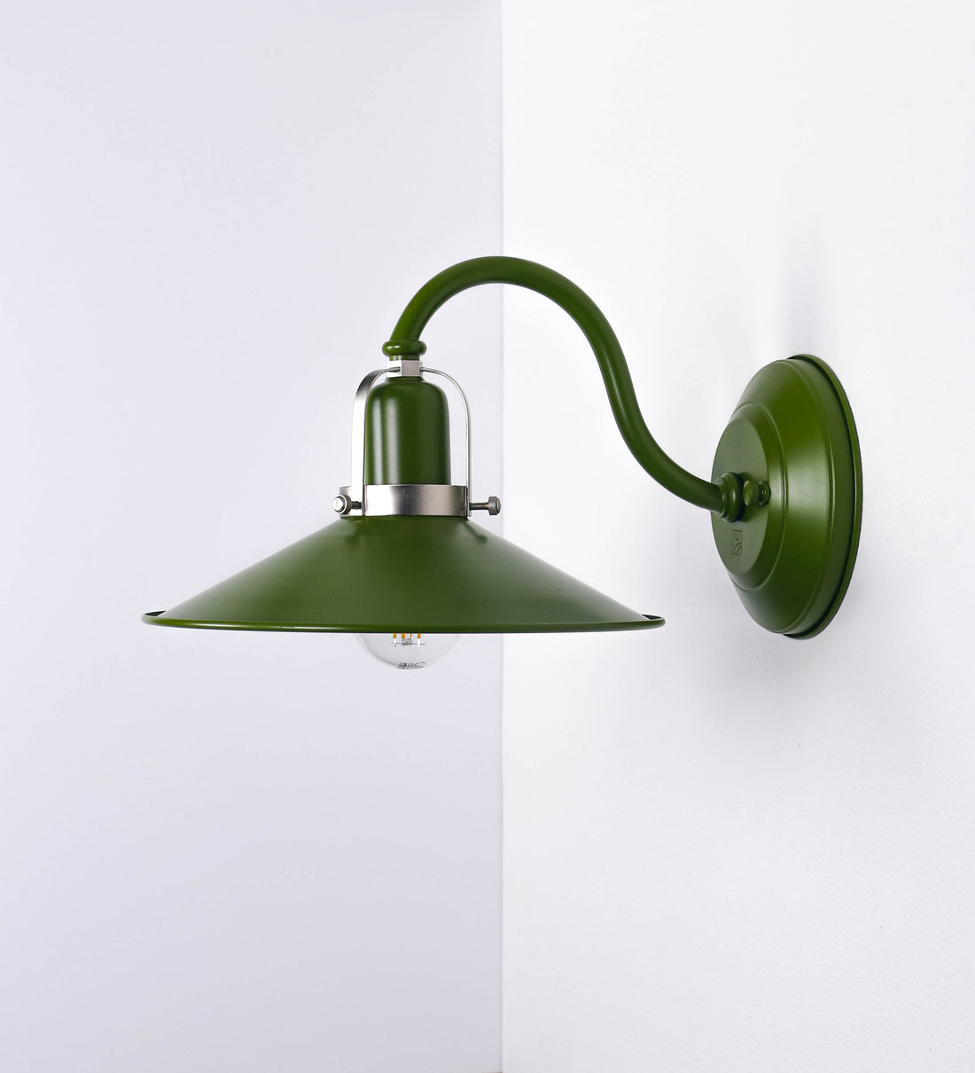 Aplique de parede com um design clássico e cores contemporâneas. O aplique é composto por uma base circular verde couve-de-bruxelas, de onde se estende um braço curvo, que forma um arco gracioso e sustenta o chapéu do candeeiro, verde também. O chapéu tem um formato cónico invertido, com uma borda ligeiramente levantada, que direciona a luz para baixo. O interior do chapéu é branco. No topo do chapéu, há um suporte circular em níquel escovado, com três braços que fixa o chapéu ao braço curvo.