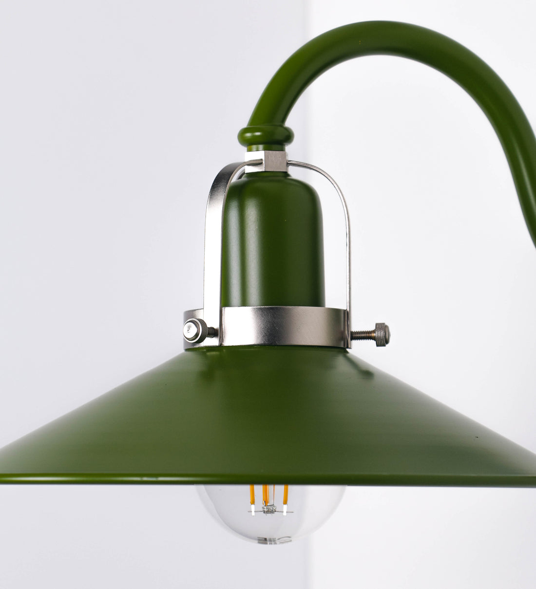 Aplique de parede com um design clássico e cores contemporâneas. O aplique é composto por uma base circular verde couve-de-bruxelas, de onde se estende um braço curvo, que forma um arco gracioso e sustenta o chapéu do candeeiro, verde também. O chapéu tem um formato cónico invertido, com uma borda ligeiramente levantada, que direciona a luz para baixo. O interior do chapéu é branco. No topo do chapéu, há um suporte circular em níquel escovado, com três braços que fixa o chapéu ao braço curvo.
