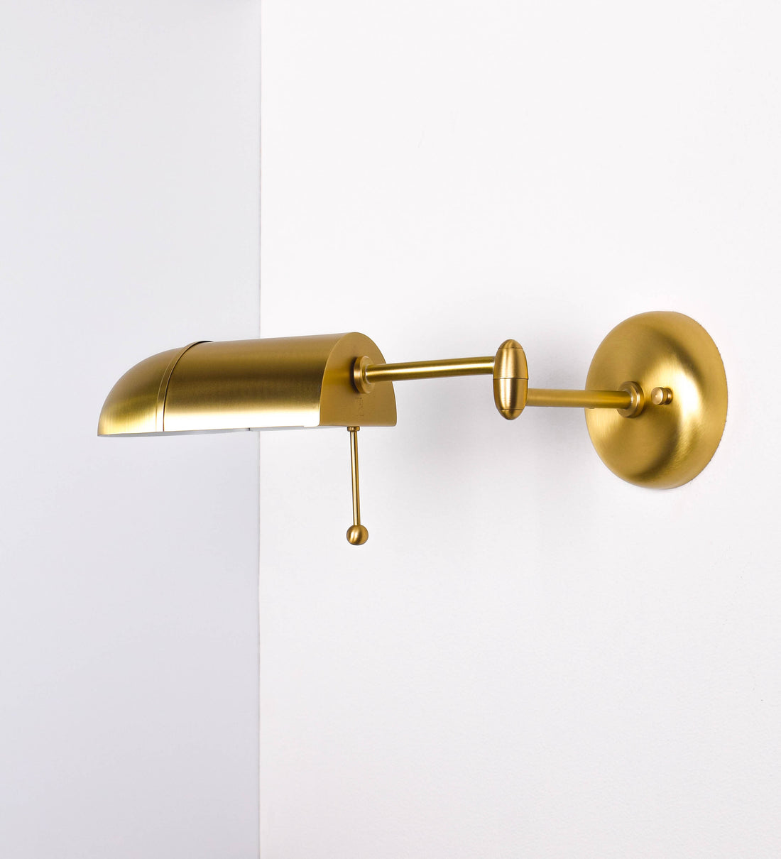 Aplique de parede com um design elegante e funcional. O aplique é todo em latão escovado. A base do aplique é redonda e fixa-se diretamente na parede. Do centro da sua base sai o braço do aplique, que é reto e horizontal. O braço é ajustável permitindo direcionar a luz. No final do braço, há um chapéu longo e arredondado e com uma pequena haste com uma esfera na ponta, que auxilia a movimentar o chapéu. O interior do chapéu é branco.