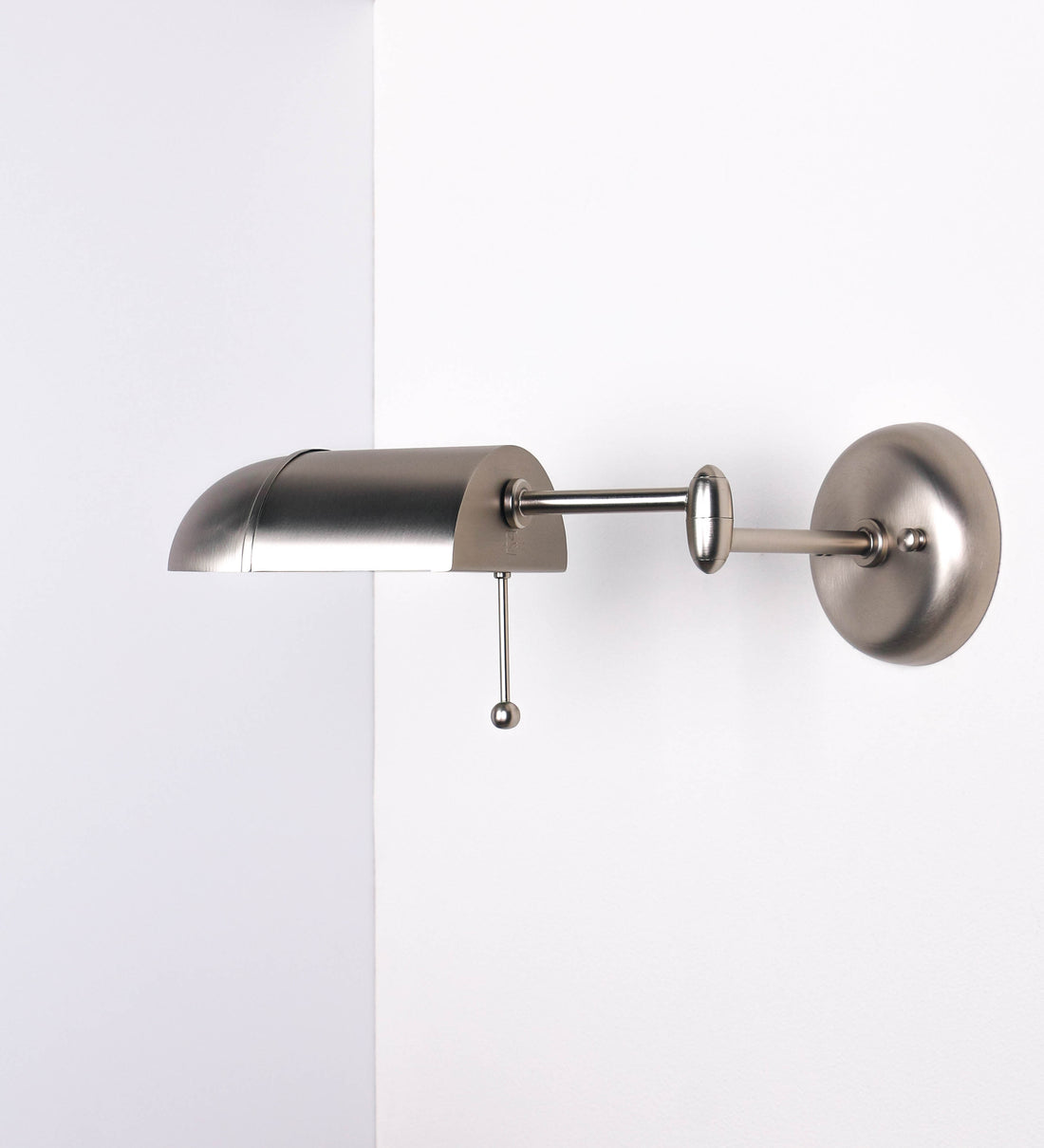 Aplique de parede com um design elegante e funcional. O aplique é todo em níquel escovado. A base do aplique é redonda e fixa-se diretamente na parede. Do centro da sua base sai o braço do aplique, que é reto e horizontal. O braço é ajustável permitindo direcionar a luz. No final do braço, há um chapéu longo e arredondado e com uma pequena haste com uma esfera na ponta, que auxilia a movimentar o chapéu. O interior do chapéu é branco.