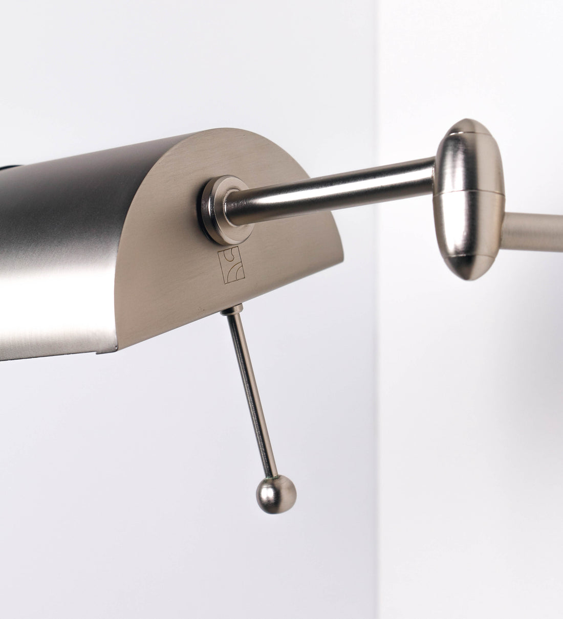 Aplique de parede com um design elegante e funcional. O aplique é todo em níquel escovado. A base do aplique é redonda e fixa-se diretamente na parede. Do centro da sua base sai o braço do aplique, que é reto e horizontal. O braço é ajustável permitindo direcionar a luz. No final do braço, há um chapéu longo e arredondado e com uma pequena haste com uma esfera na ponta, que auxilia a movimentar o chapéu. O interior do chapéu é branco.