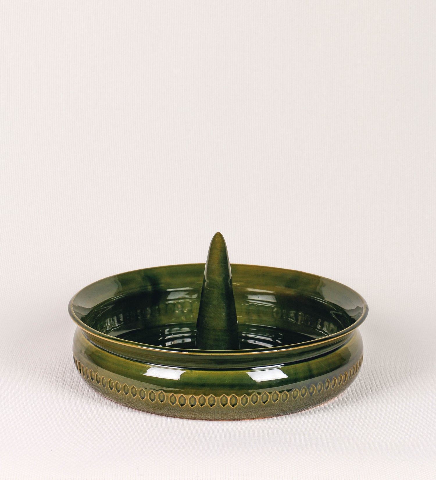 Assadeira para frangos com um design rústico e distinto. O assa-frangos é de barro, vidrado em tom verde vidro com brilho, conferindo um caráter vintage à peça. A forma do assa-frangos é circular, com uma borda superior mais estreita e um espeto cónico centrado no interior para colocar os frangos. A superfície externa da peça é decorada com um padrão de relevos losangulares, que envolvem o assa-frangos e acrescentam textura e interesse visual ao design.