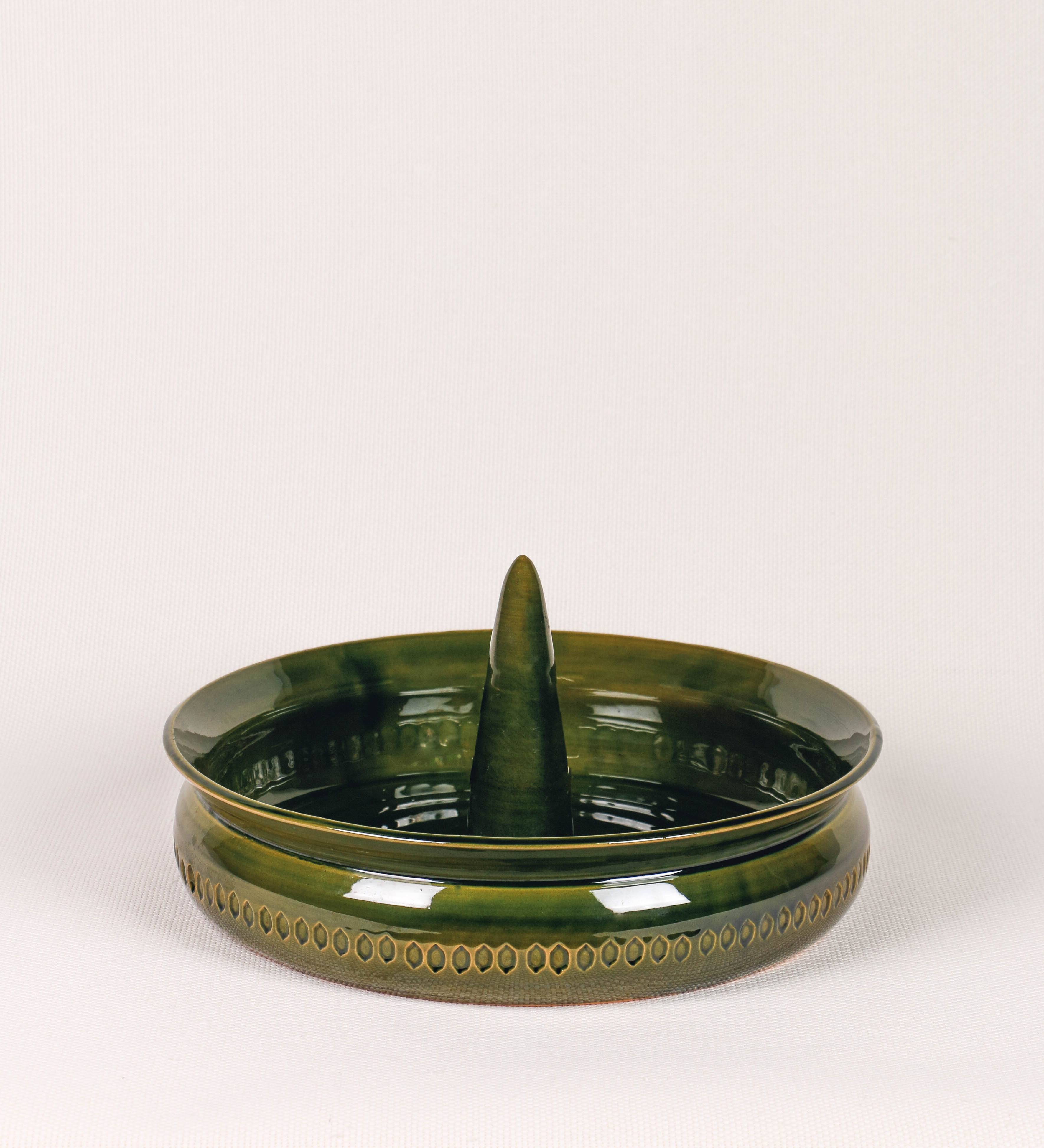 Assadeira para frangos com um design rústico e distinto. O assa-frangos é de barro, vidrado em tom verde vidro com brilho, conferindo um caráter vintage à peça. A forma do assa-frangos é circular, com uma borda superior mais estreita e um espeto cónico centrado no interior para colocar os frangos. A superfície externa da peça é decorada com um padrão de relevos losangulares, que envolvem o assa-frangos e acrescentam textura e interesse visual ao design.