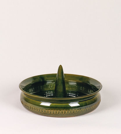 Assadeira para frangos com um design rústico e distinto. O assa-frangos é de barro, vidrado em tom verde vidro com brilho, conferindo um caráter vintage à peça. A forma do assa-frangos é circular, com uma borda superior mais estreita e um espeto cónico centrado no interior para colocar os frangos. A superfície externa da peça é decorada com um padrão de relevos losangulares, que envolvem o assa-frangos e acrescentam textura e interesse visual ao design.