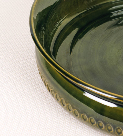 Assadeira para frangos com um design rústico e distinto. O assa-frangos é de barro, vidrado em tom verde vidro com brilho, conferindo um caráter vintage à peça. A forma do assa-frangos é circular, com uma borda superior mais estreita e um espeto cónico centrado no interior para colocar os frangos. A superfície externa da peça é decorada com um padrão de relevos losangulares, que envolvem o assa-frangos e acrescentam textura e interesse visual ao design.