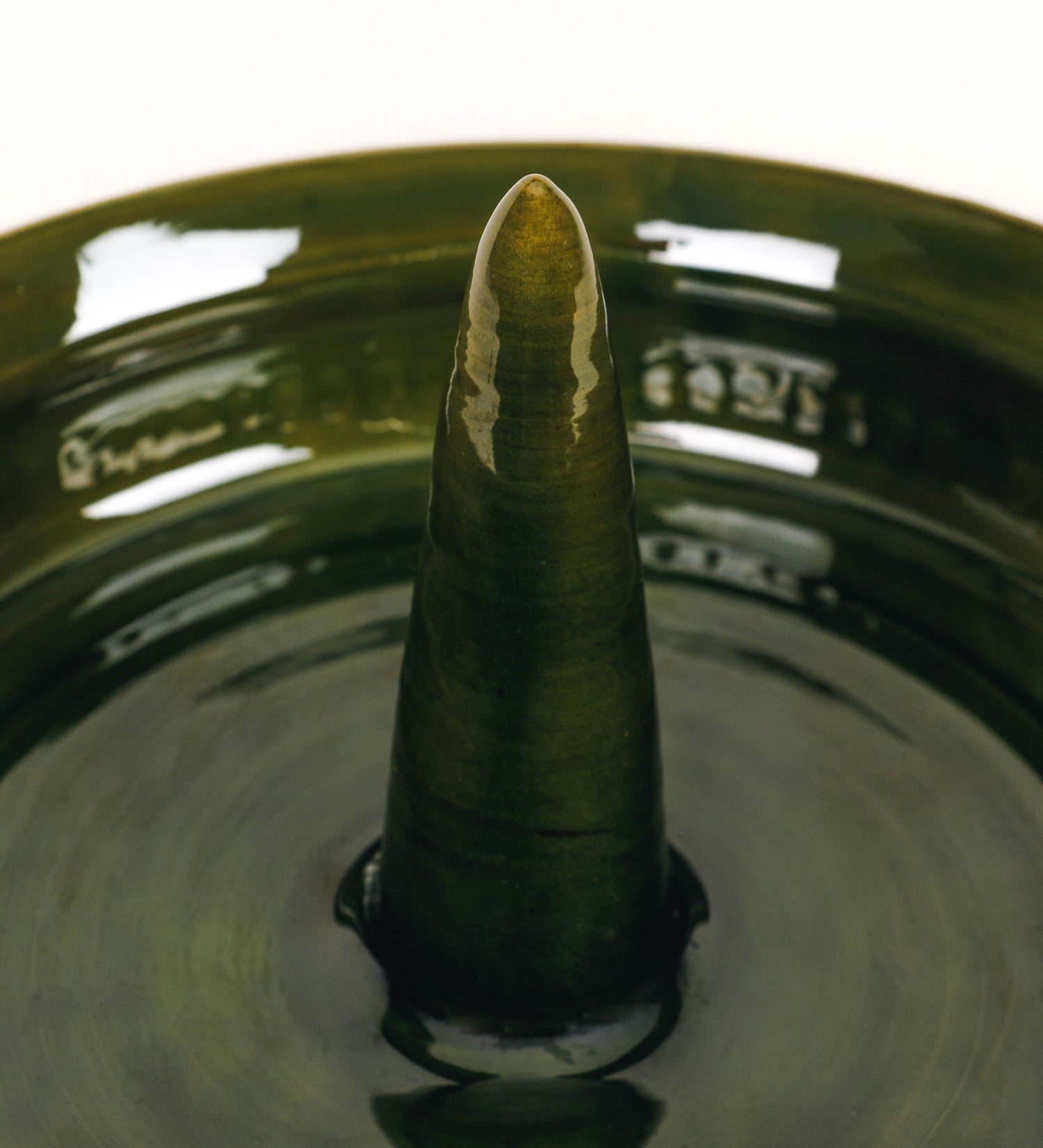 Assadeira para frangos com um design rústico e distinto. O assa-frangos é de barro, vidrado em tom verde vidro com brilho, conferindo um caráter vintage à peça. A forma do assa-frangos é circular, com uma borda superior mais estreita e um espeto cónico centrado no interior para colocar os frangos. A superfície externa da peça é decorada com um padrão de relevos losangulares, que envolvem o assa-frangos e acrescentam textura e interesse visual ao design.